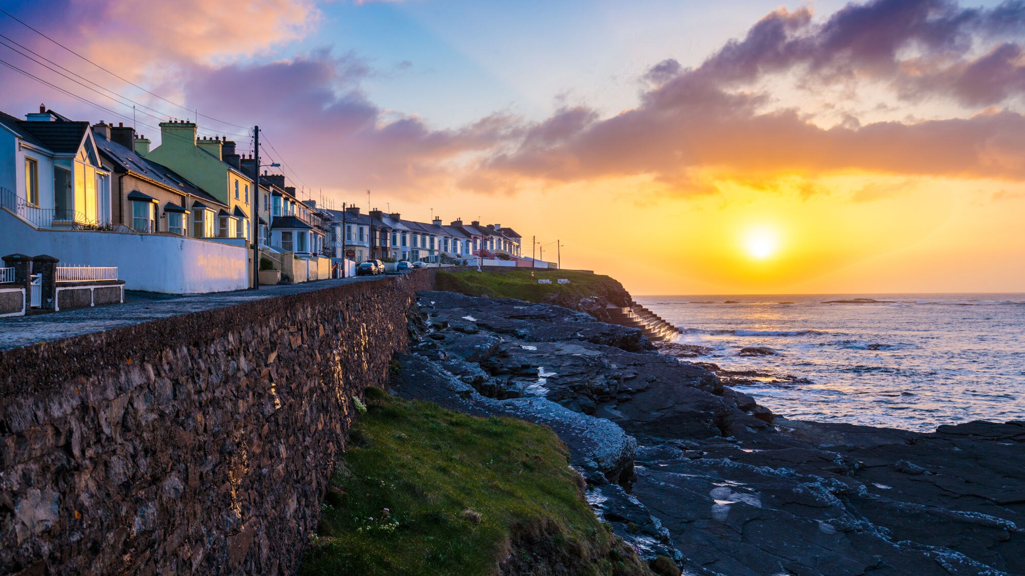 Sonnenuntergang über der Küste von Kilkee mit Reihenhäusern auf einer Steinmauer und Felsen am Meer