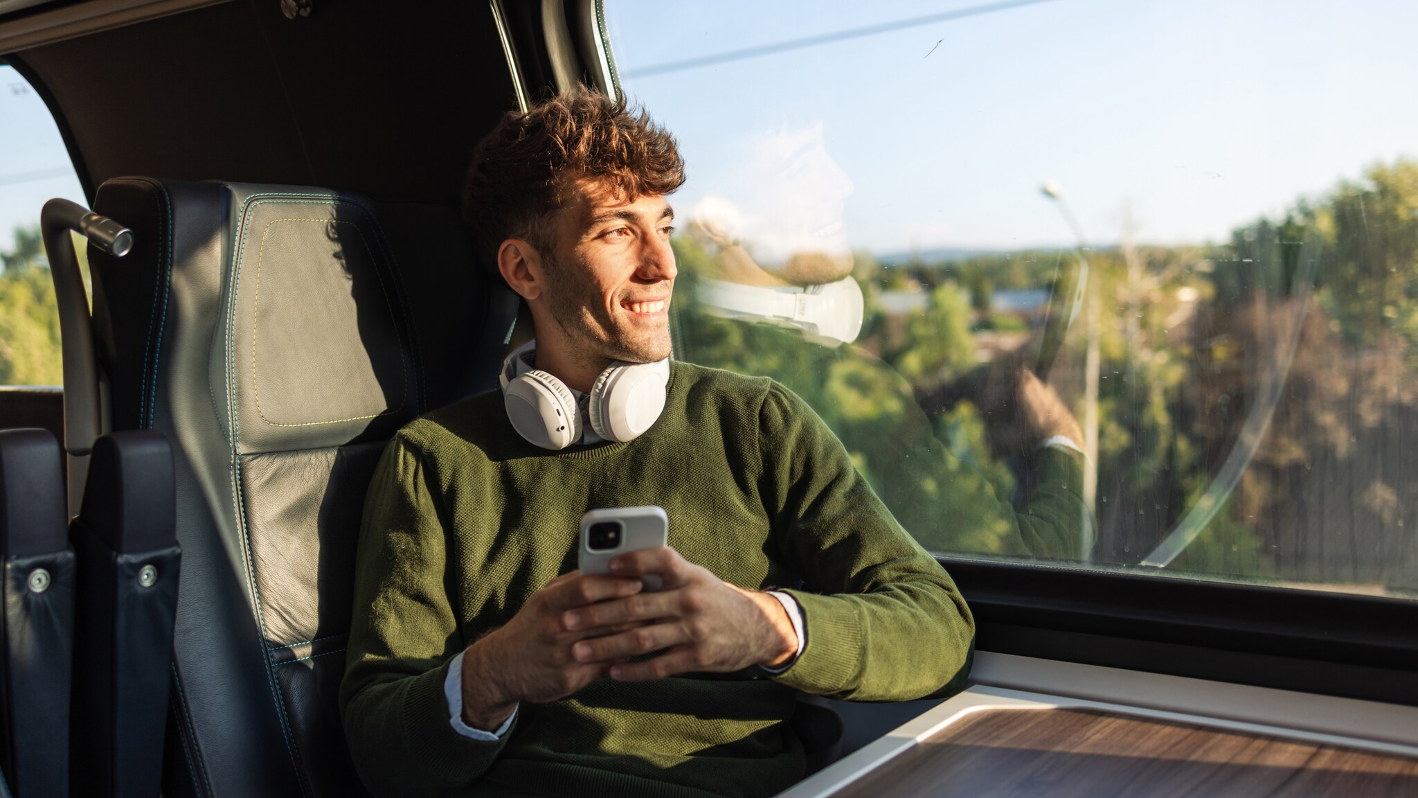 Junger Mann mit grünem Pullover und weißen Kopfhörern sitzt im Zug mit Smartphone in der Hand und blickt aus dem Fenster.