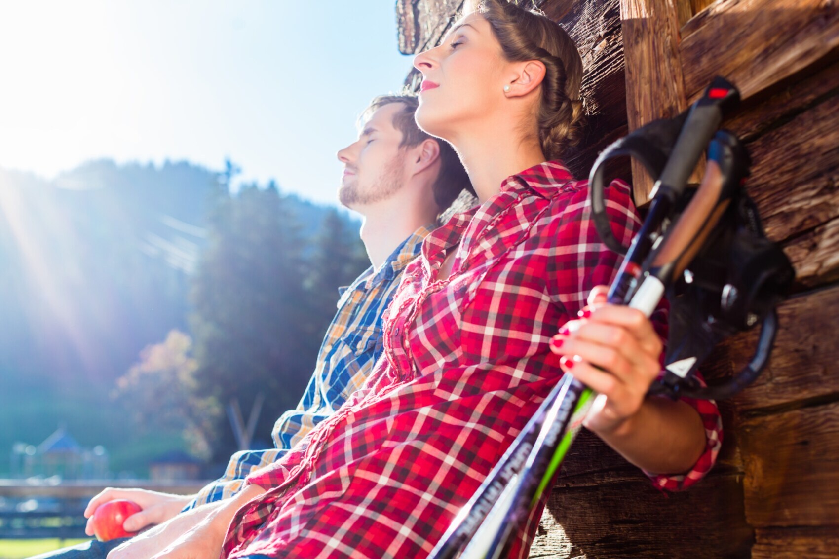 Junges Paar ruht sich nach Bergwanderung mit Trekking-Stöcken in der Hand an eine Holzhütte gelehnt aus