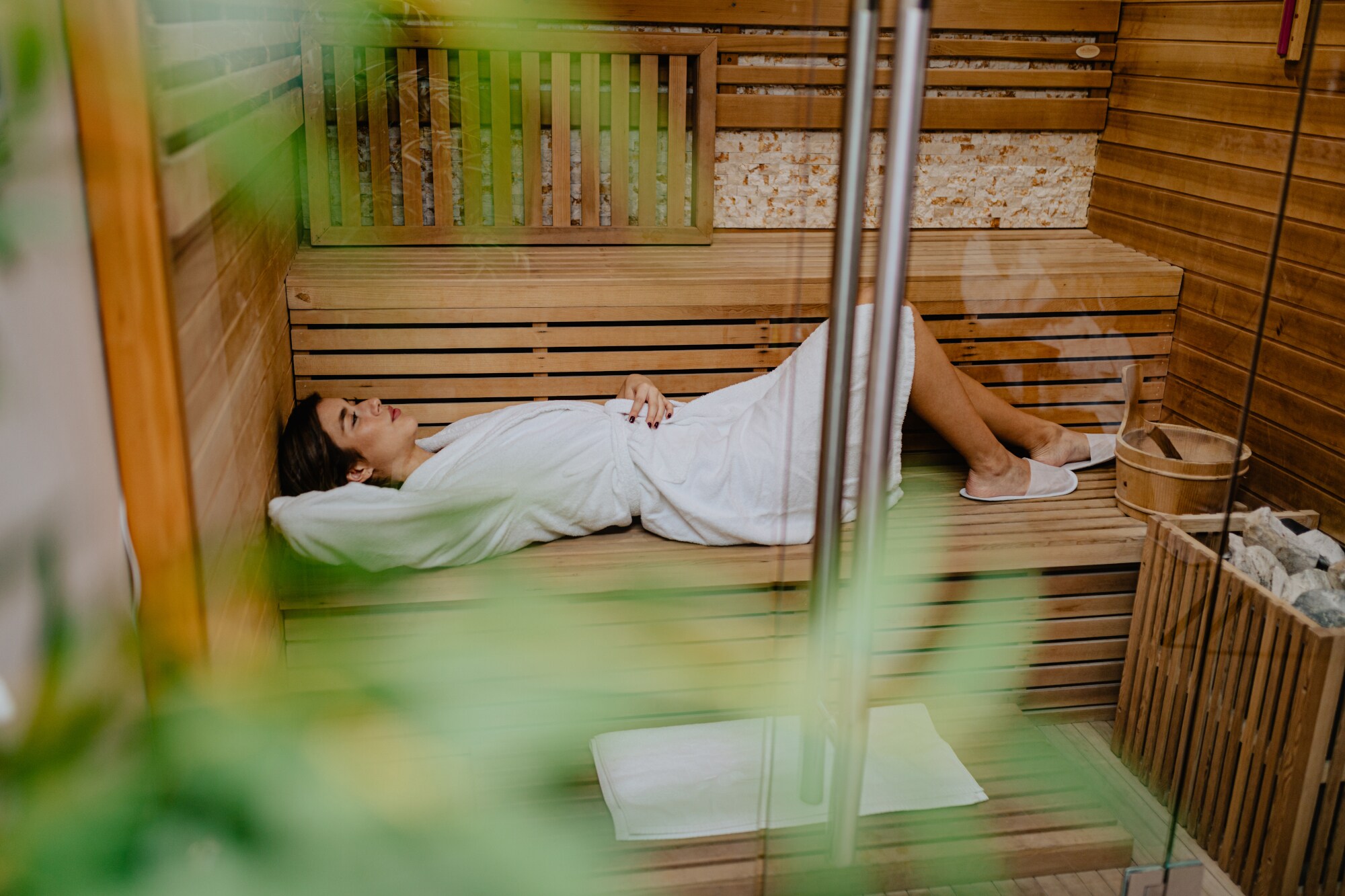 Blick durch eine Glastür auf eine junge Frau, die im Bademantel entspannt in einer Holzsauna liegt