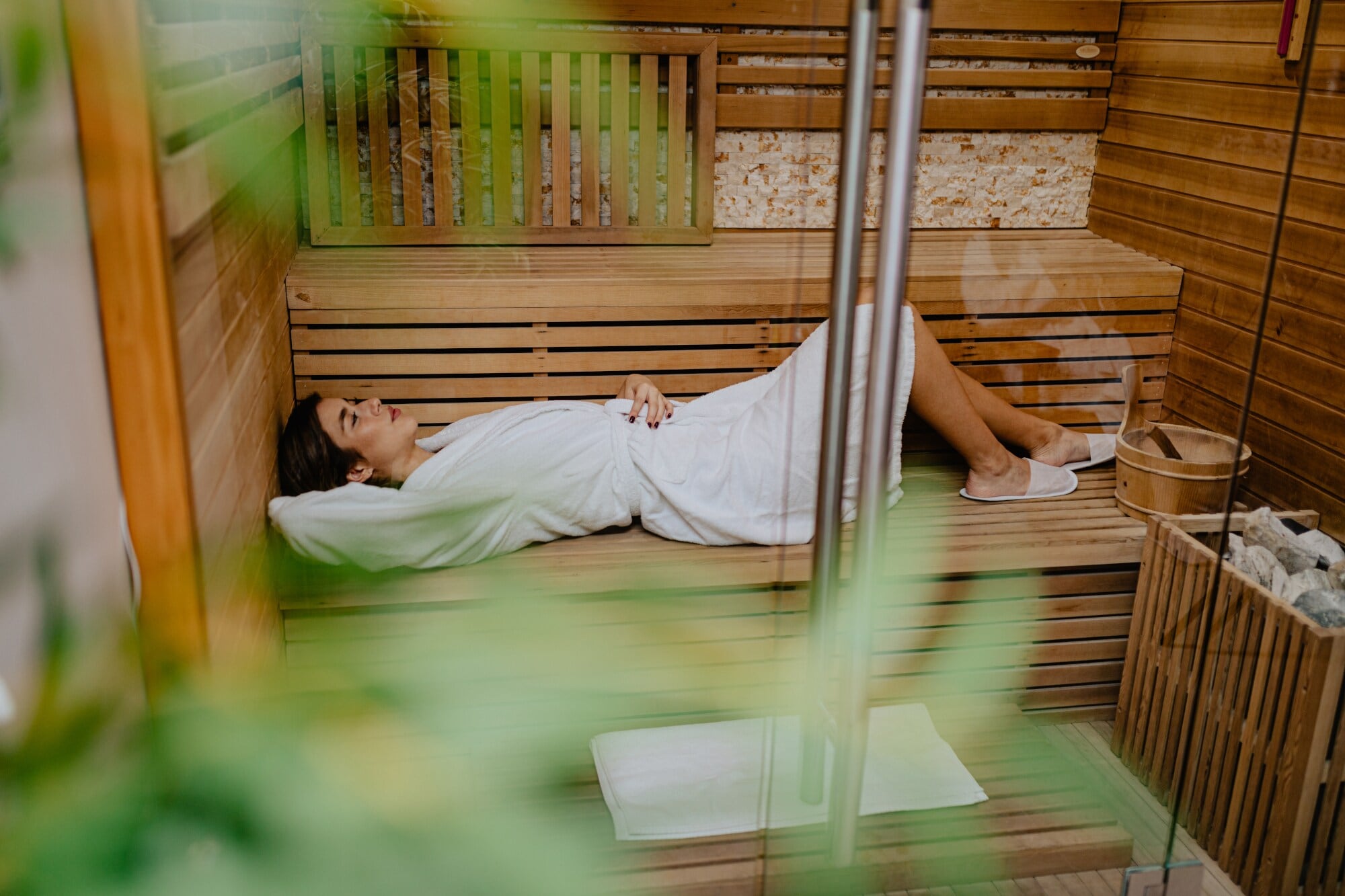 Blick durch eine Glastür auf eine junge Frau, die im Bademantel entspannt in einer Holzsauna liegt Blick durch eine Glastür auf eine junge Frau, die im Bademantel entspannt in einer Holzsauna liegt