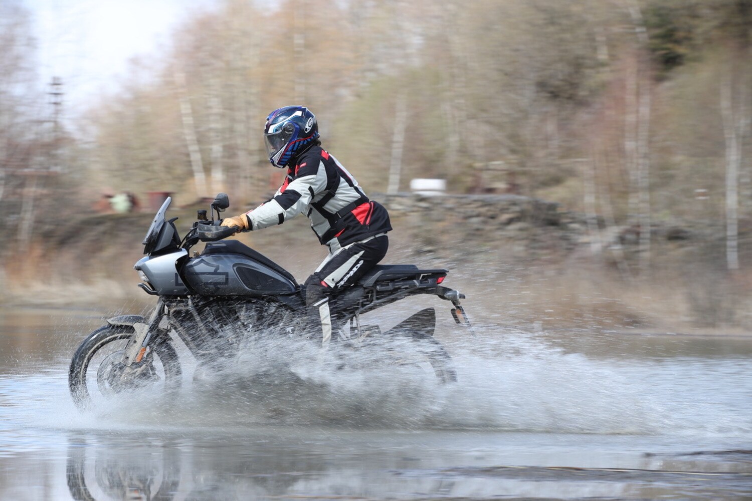 Ein Motorradfahrer in Endurobekleidung fährt auf einer grauen Harley-Davidson durch ein Flussbett und lässt Wasser aufspritzen.