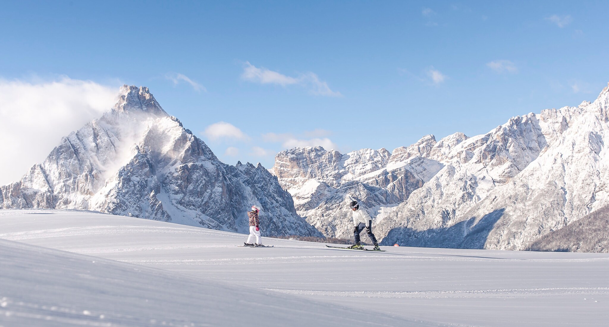 Ein Erwachsener und ein Kind auf Skiern auf einer Piste vor den schneebedeckten Gipfeln eines Bergmassivs