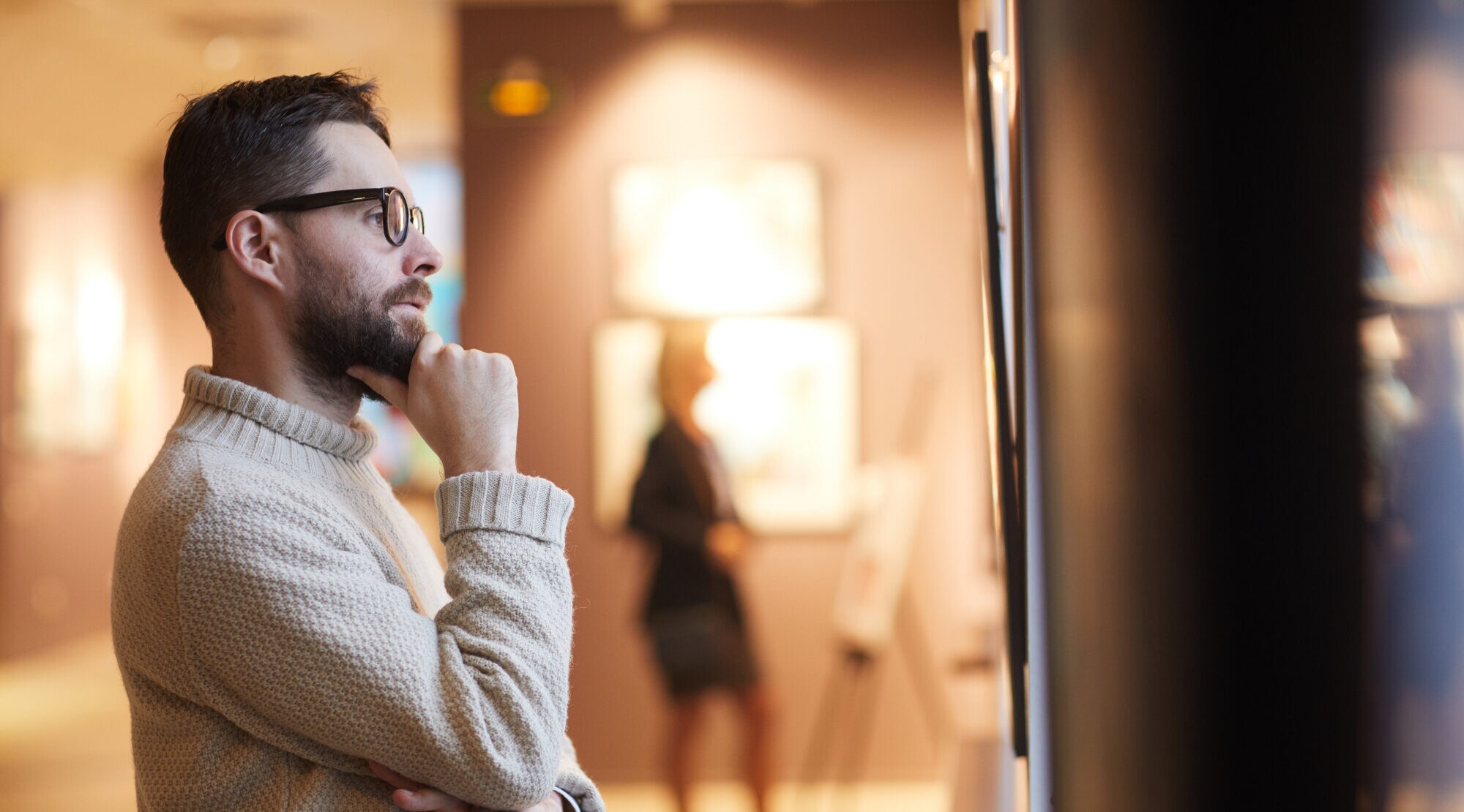 Ein Mann mit Brille und Rollkragenpullover betrachtet interessiert ein Kunstwerk in einer Galerie