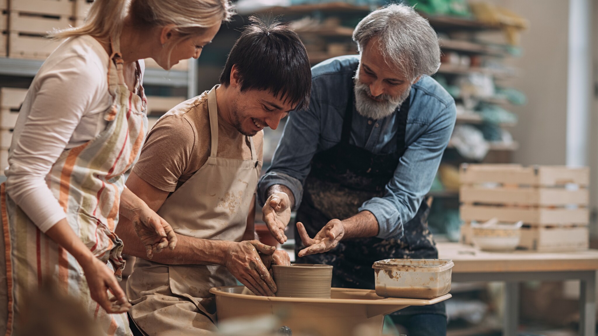 Drei Personen in einer Töpferwerkstatt formen gemeinsam Ton an einer Töpferscheibe, alle tragen Schürzen und lächeln. Drei Personen in einer Töpferwerkstatt formen gemeinsam Ton an einer Töpferscheibe, alle tragen Schürzen und lächeln.