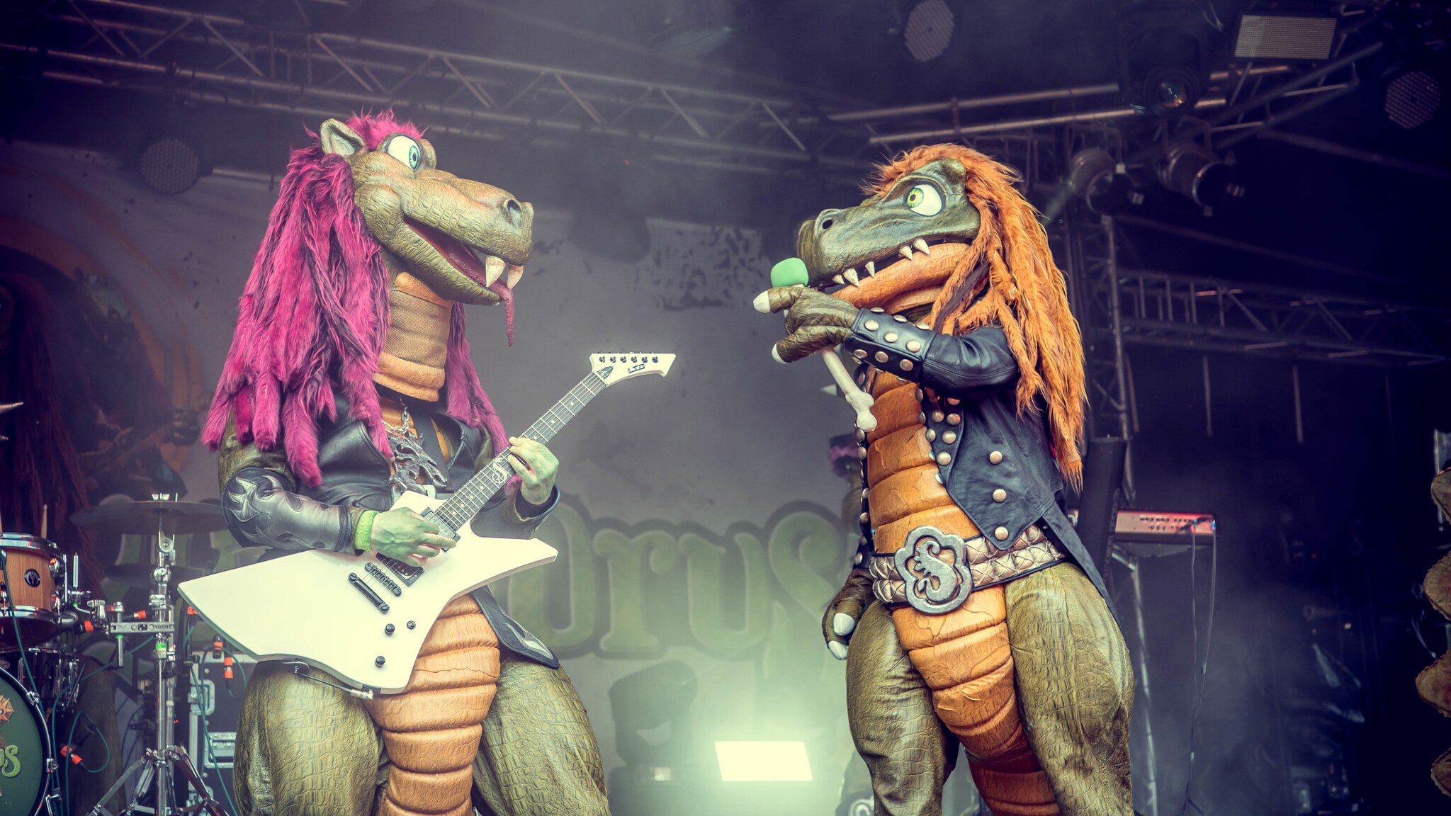 Eine Person im Drachenkostüm, die eine Gitarre hält und eine Person im Dinosaurierkostüm mit Mikrofon ansieht