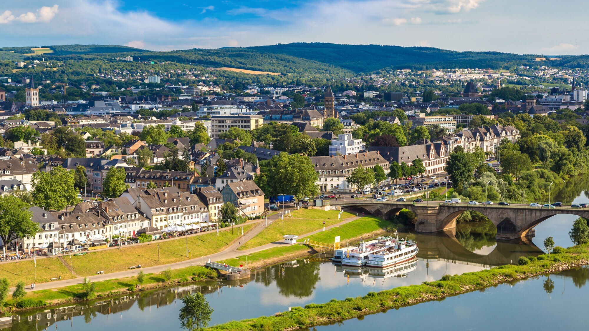 Blick auf Trier mit Mosel, historischen Gebäuden, Brücke und bewaldeten Hügeln im Hintergrund bei sonnigem Wetter.