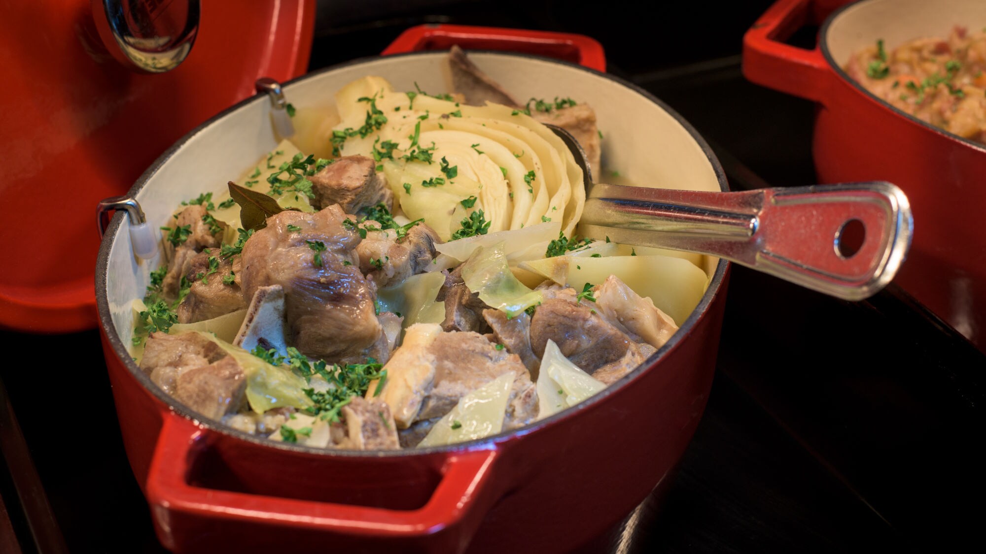 Roter Bräter mit Fårikål, bestehend aus Lammfleisch, Kohl und Kartoffeln, garniert mit Petersilie, daneben ein weiterer roter Topf mit Reisgericht. Roter Bräter mit Fårikål, bestehend aus Lammfleisch, Kohl und Kartoffeln, garniert mit Petersilie, daneben ein weiterer roter Topf mit Reisgericht.