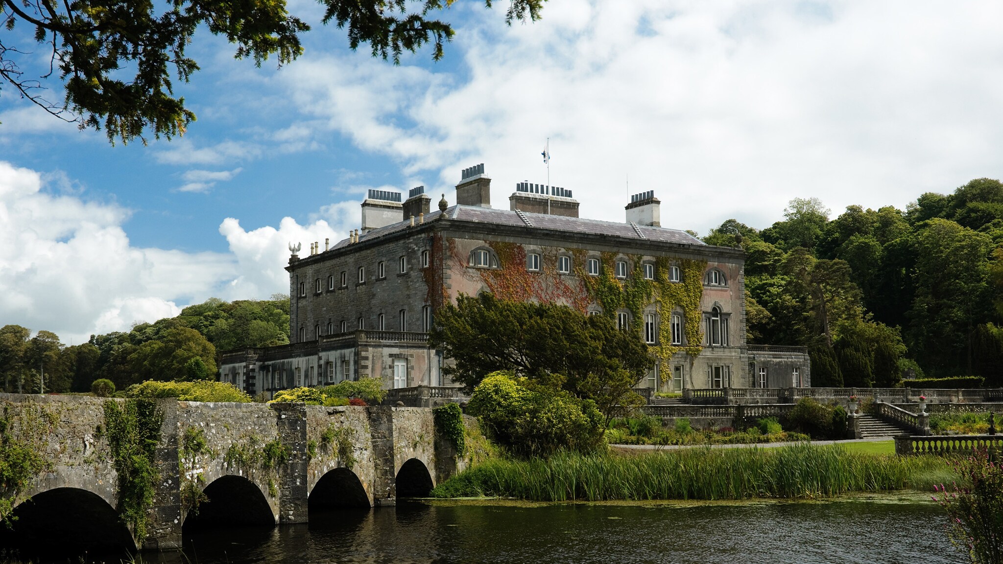 Vor einem großen, rechteckigen Haus mit vielen kleinen Fenstern liegt ein See mit einer stabilen Steinbrücke.