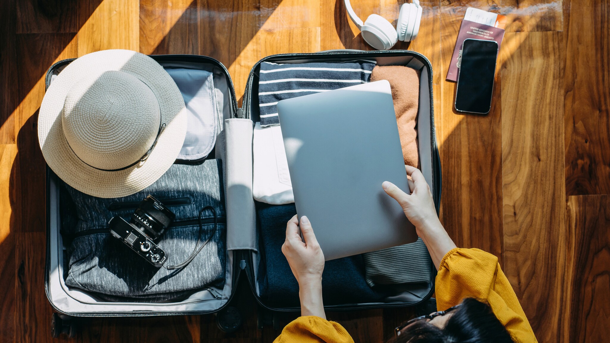 Offener Koffer mit Sonnenhut, Kamera, Kleidung, Laptop, weißen Kopfhörern und Smartphone auf Holzboden, Person legt Laptop in Koffer.