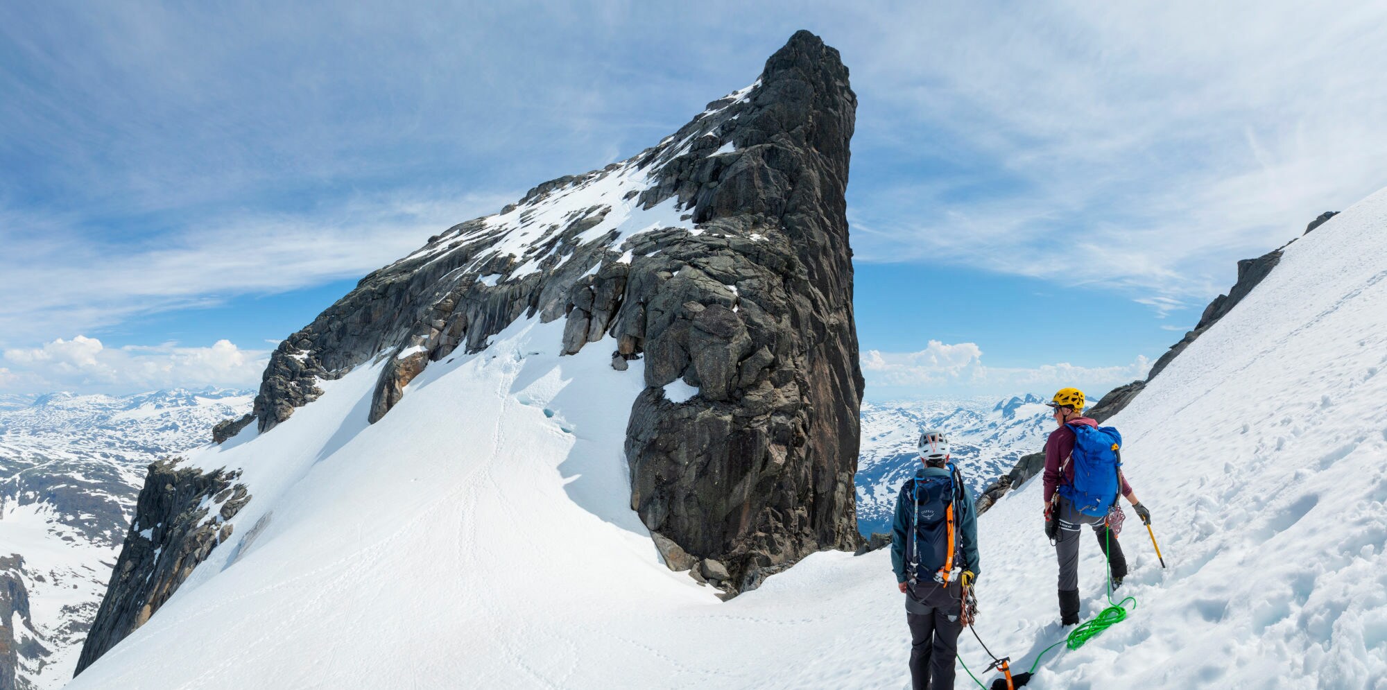Zwei Bergsteiger mit Helmen und Rucksäcken stehen auf schneebedecktem Grat in Jotunheimen, Blick auf steilen Felsgipfel und umliegende Berglandschaft. Zwei Bergsteiger mit Helmen und Rucksäcken stehen auf schneebedecktem Grat in Jotunheimen, Blick auf steilen Felsgipfel und umliegende Berglandschaft.