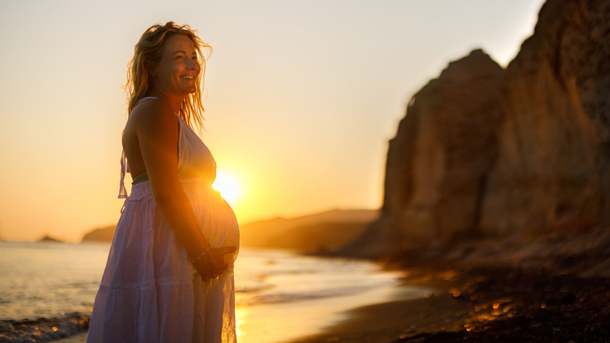 Eine lächelnde schwangere Frau im weißen Sommerkleid hält sich ihren Bauch an einem Strand mit Felsen bei Sonnenuntergang.