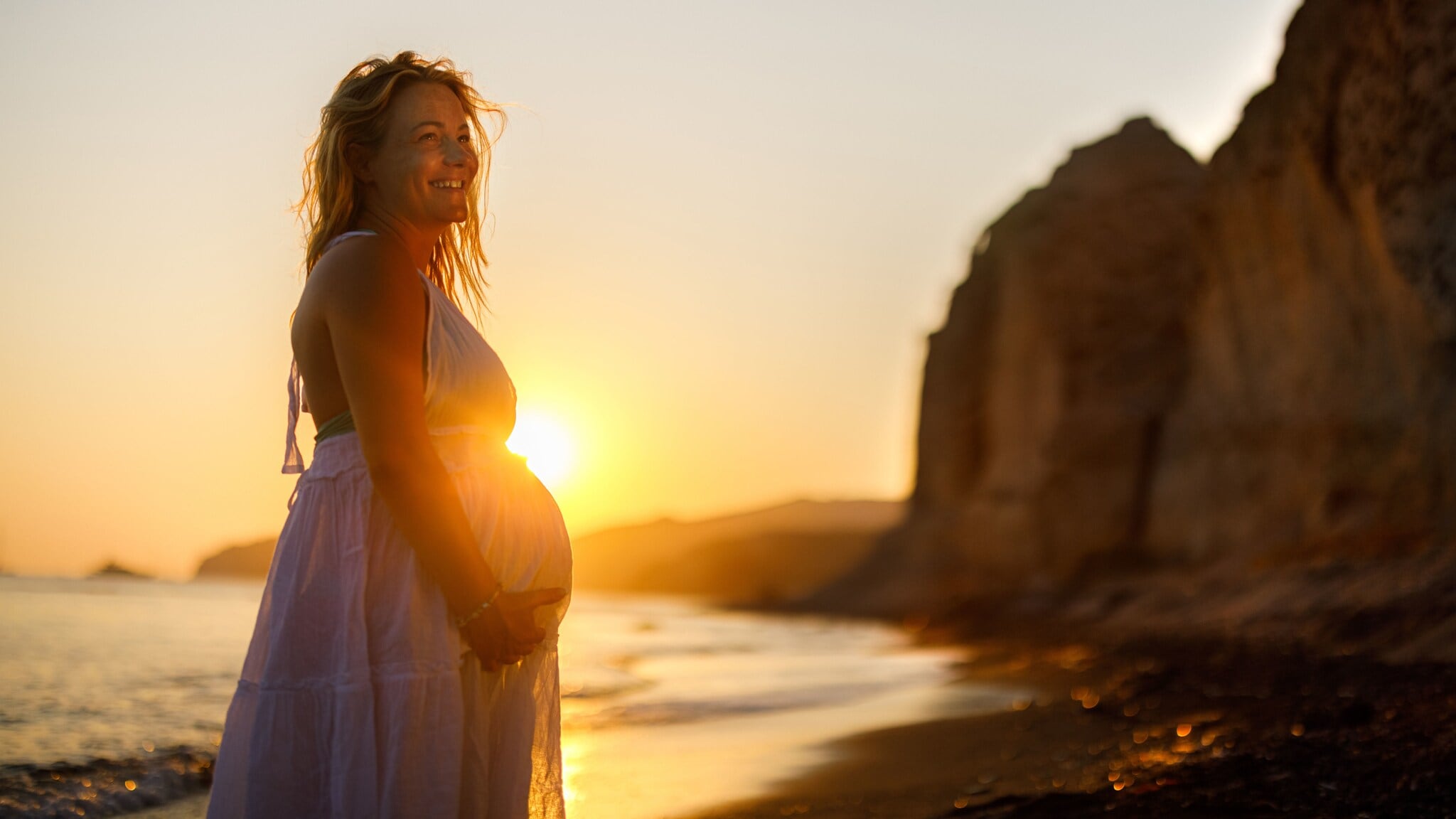Eine lächelnde schwangere Frau im weißen Sommerkleid hält sich ihren Bauch an einem Strand mit Felsen bei Sonnenuntergang. Eine lächelnde schwangere Frau im weißen Sommerkleid hält sich ihren Bauch an einem Strand mit Felsen bei Sonnenuntergang.