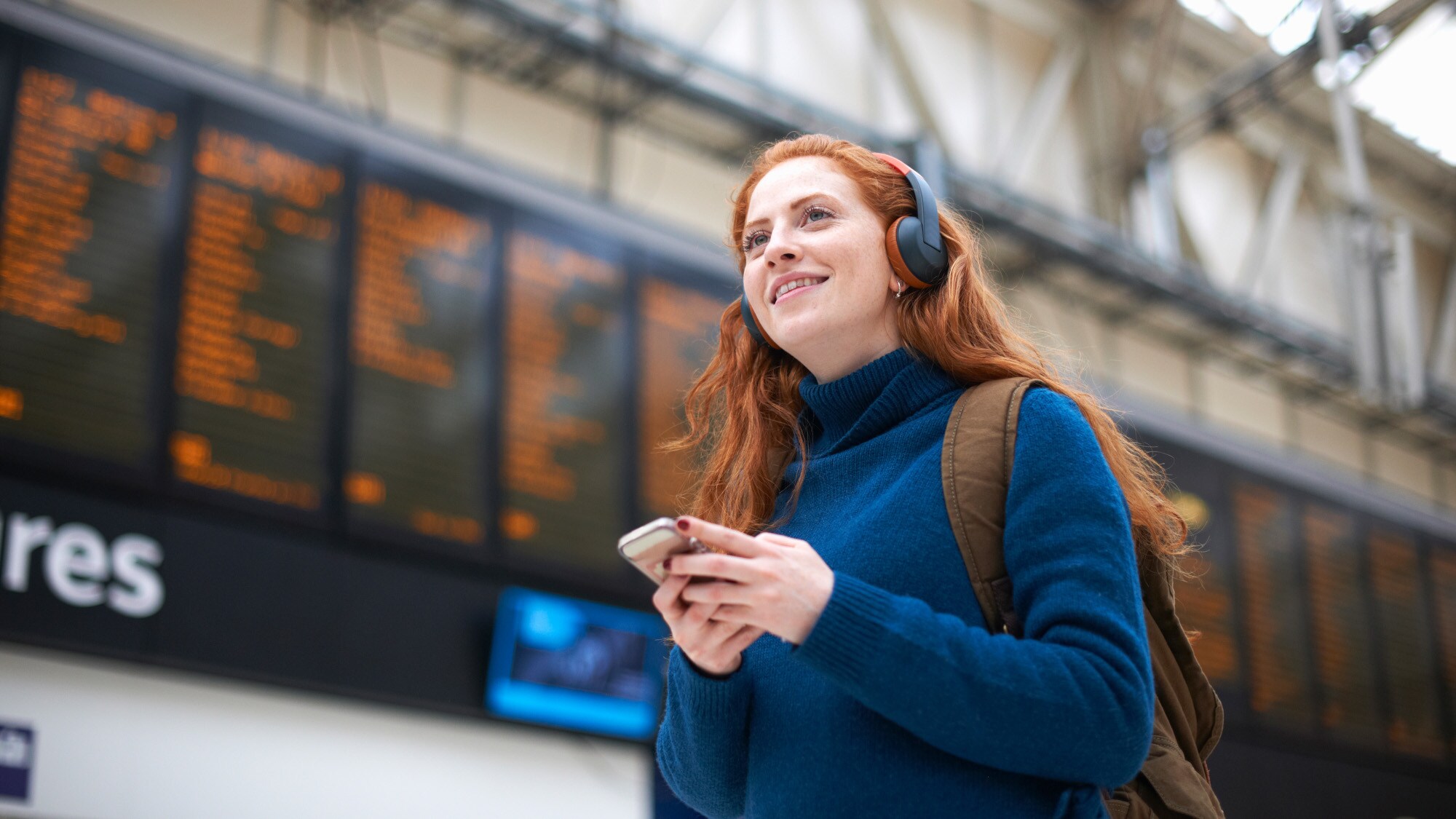 Eine junge Frau mit Kopfhörern und Smartphone steht lächelnd vor einer Abflugtafel in einem Flughafenterminal.