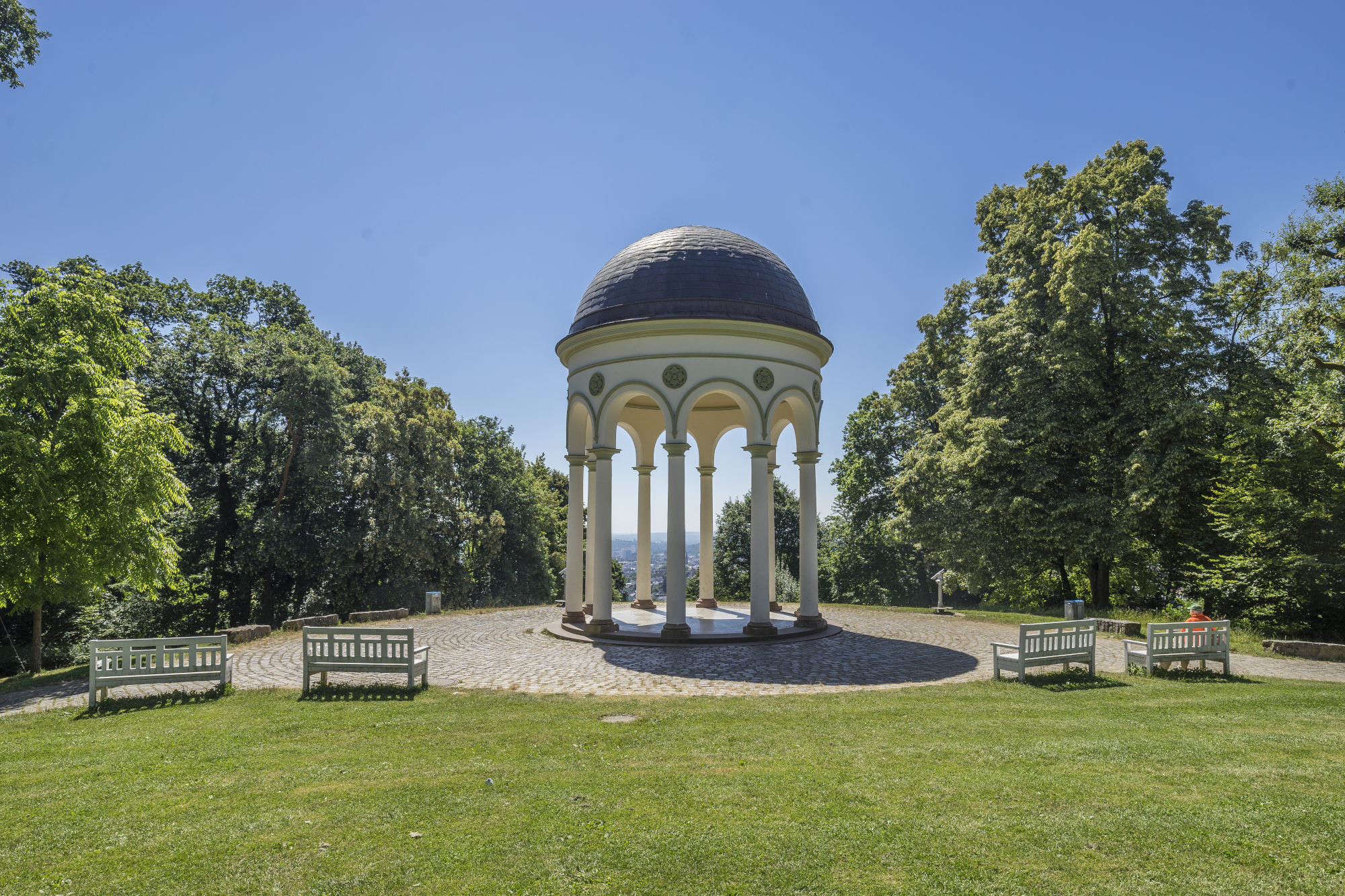 Charmanter historischer Pavillon mit Säulen und Runddach auf einer Aussichtsplattform mit Blick auf die Umgebung und gesäumt von weißen Bänken, Bäumen und einer Rasenfläche. Charmanter historischer Pavillon mit Säulen und Runddach auf einer Aussichtsplattform mit Blick auf die Umgebung und gesäumt von weißen Bänken, Bäumen und einer Rasenfläche.