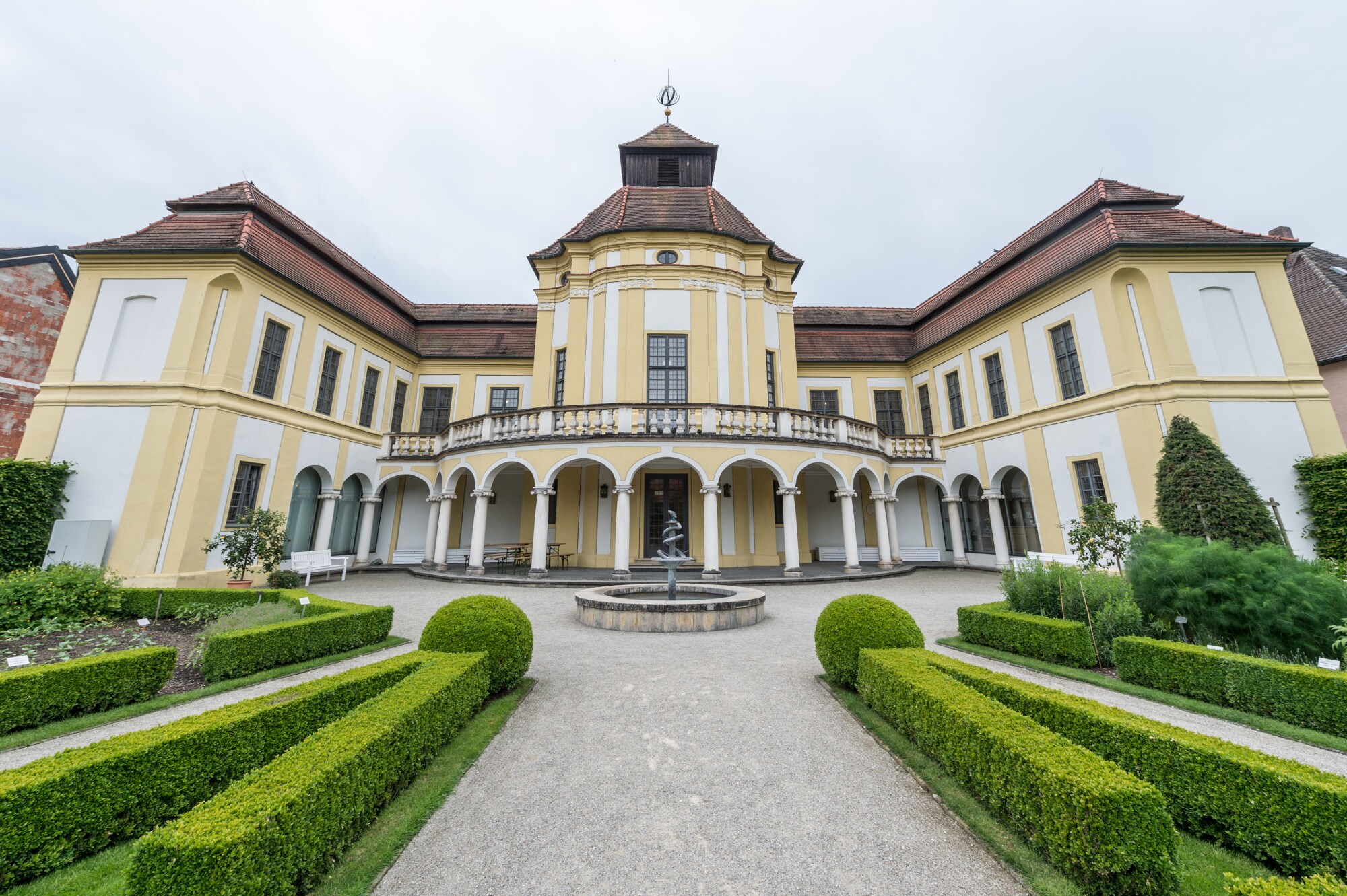 Das prächtige Gebäude des Deutschen Medizinhistorischen Museums mit weiß-gelber Fassade, davor Begrünung und ein Springbrunnen mit Äskulapstab Das prächtige Gebäude des Deutschen Medizinhistorischen Museums mit weiß-gelber Fassade, davor Begrünung und ein Springbrunnen mit Äskulapstab