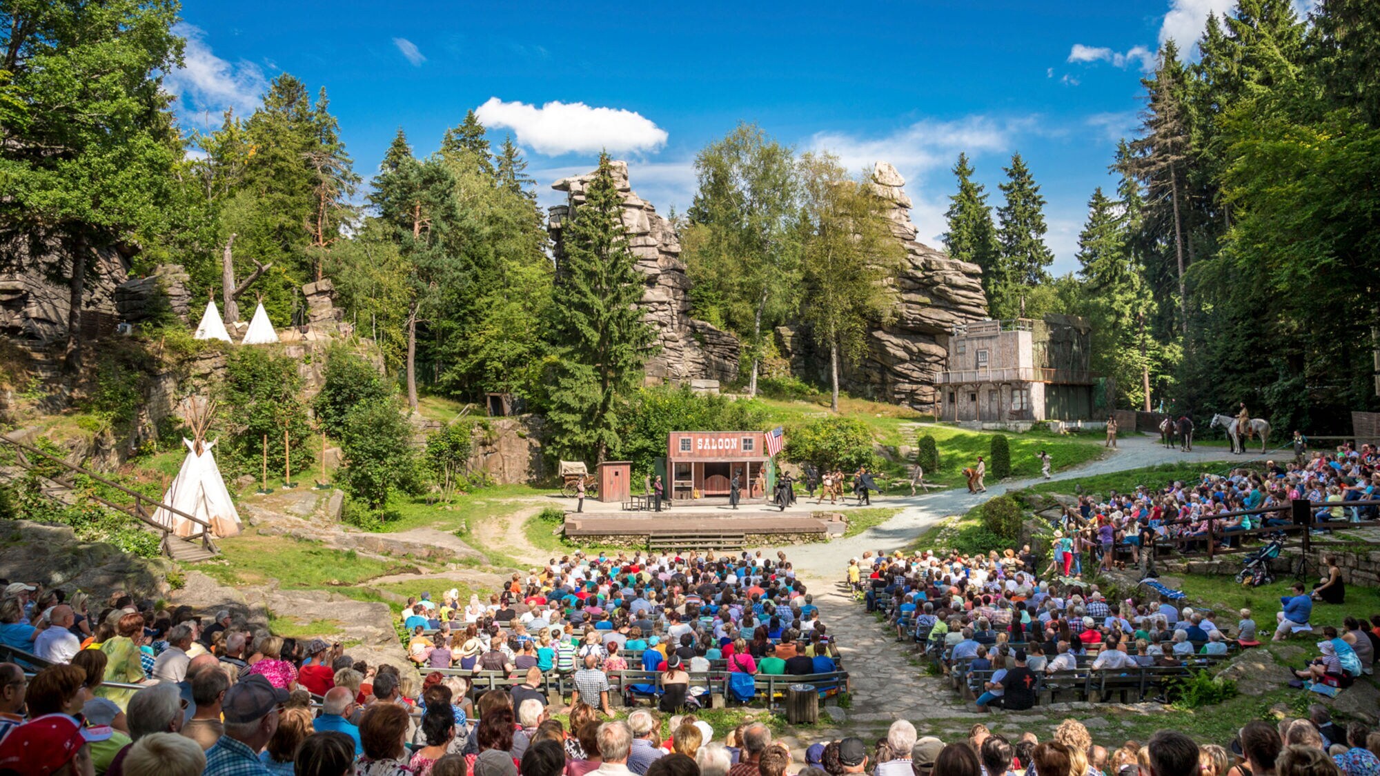 Eine Aufführung mit Publikum in einem Freilichttheater vor Waldkulisse mit Felsformationen. Eine Aufführung mit Publikum in einem Freilichttheater vor Waldkulisse mit Felsformationen.