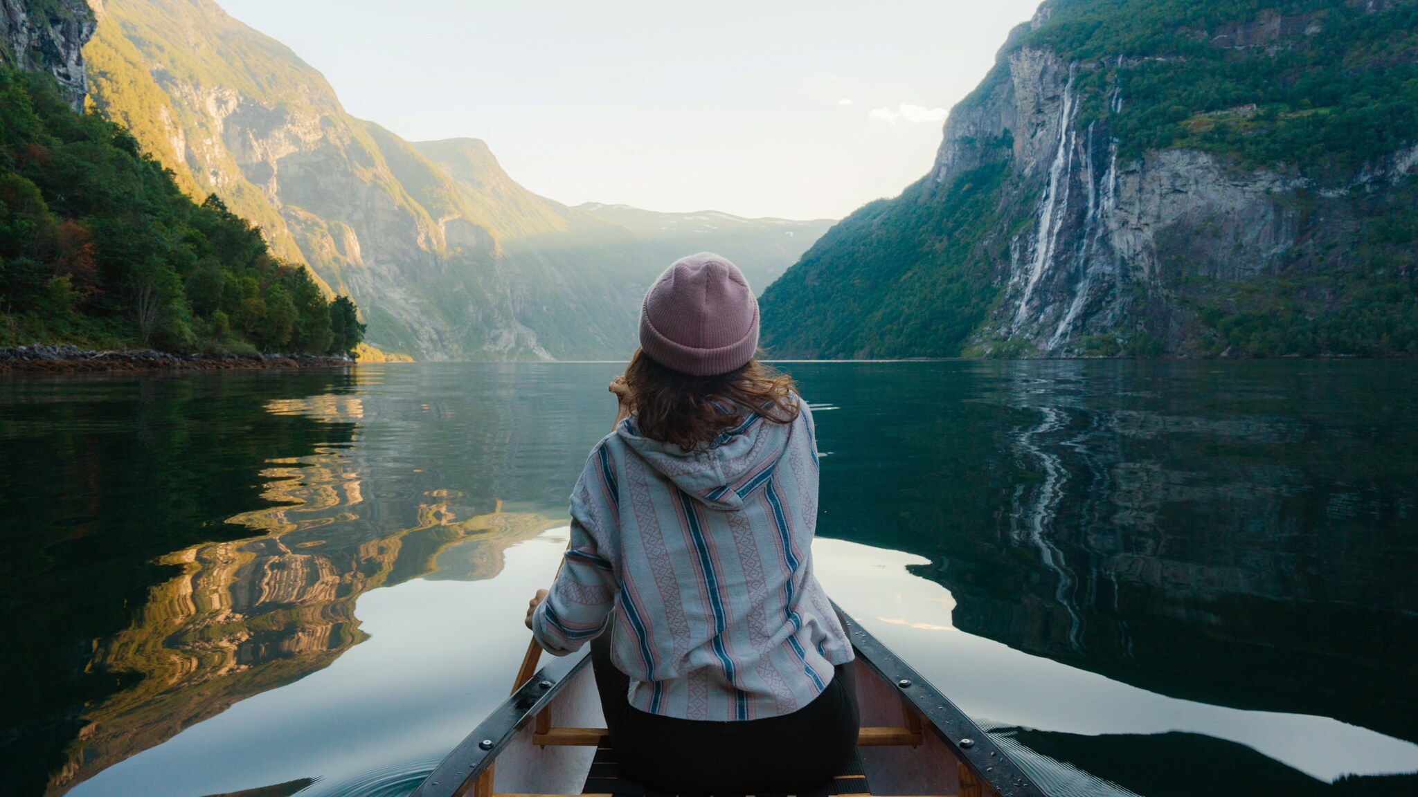Person mit rosa Mütze und gestreiftem Pullover sitzt in einem Kanu auf ruhigem Wasser, umgeben von hohen, bewaldeten norwegischen Fjordwänden mit Wasserfall. Person mit rosa Mütze und gestreiftem Pullover sitzt in einem Kanu auf ruhigem Wasser, umgeben von hohen, bewaldeten norwegischen Fjordwänden mit Wasserfall.