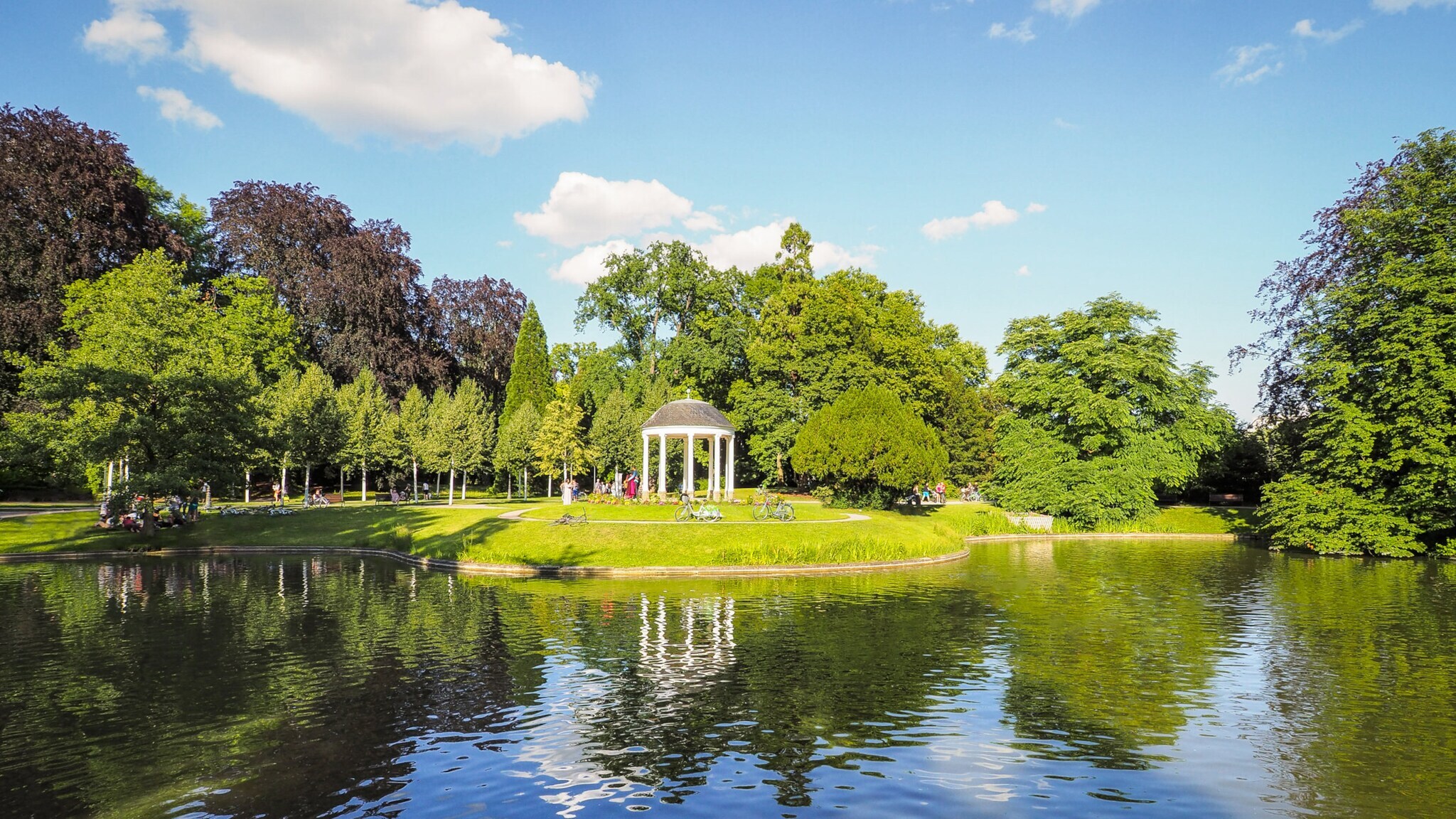 Parkanlage mit einem weißen Pavillon am Ufer eines Sees unter blauem Himmel mit Wolken Parkanlage mit einem weißen Pavillon am Ufer eines Sees unter blauem Himmel mit Wolken