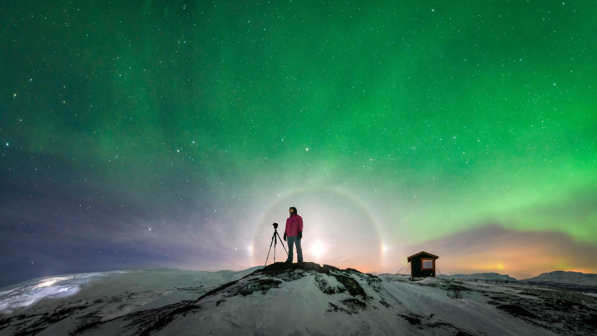 Person in roter Jacke steht auf schneebedecktem Hügel neben Kamera auf Stativ, darüber grüne Nordlichter am Nachthimmel in Schweden.