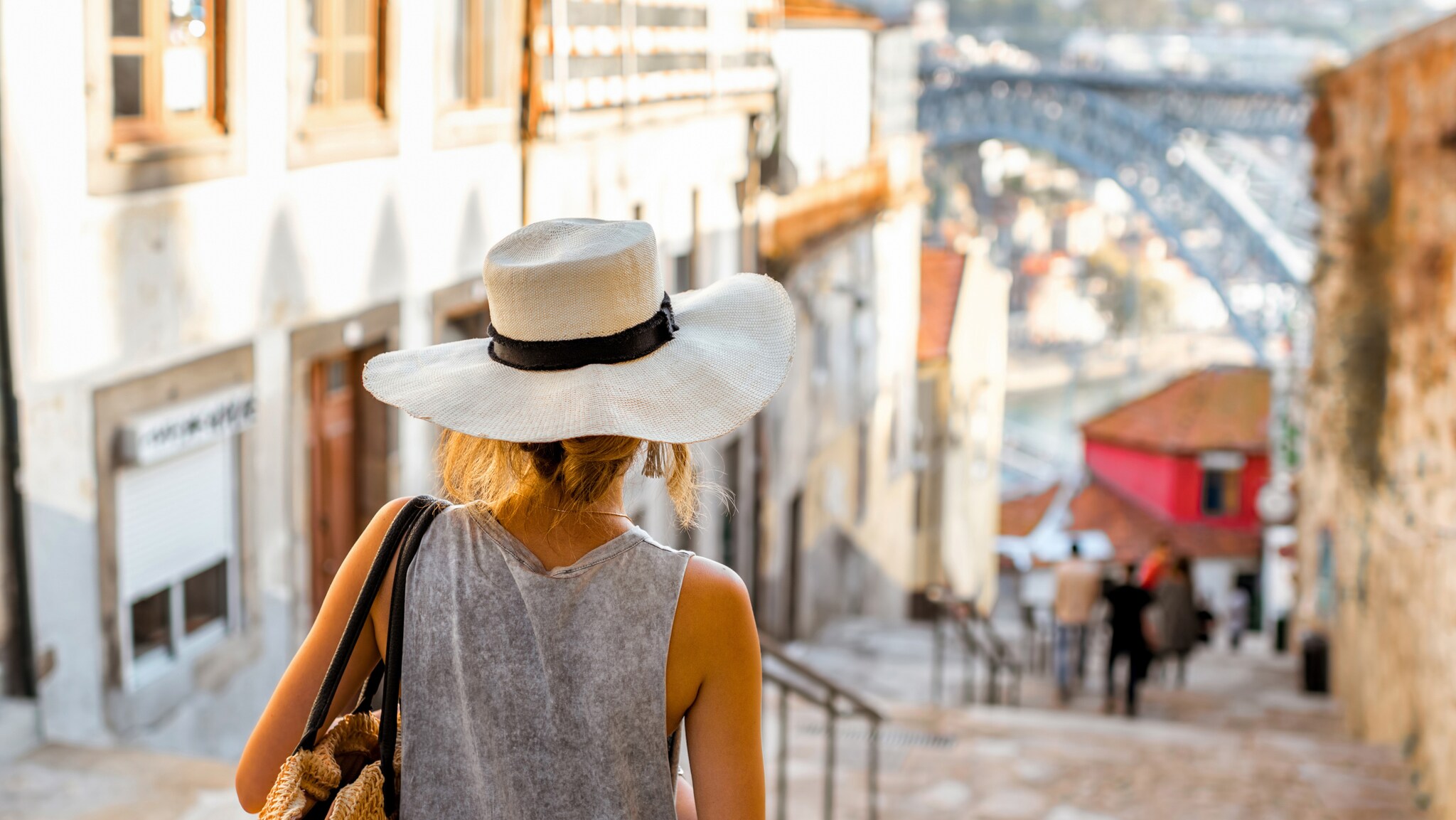 Rückansicht einer Frau mit weißem Sonnenhut, die eine Steintreppe in einer Altstadt hinuntergeht.