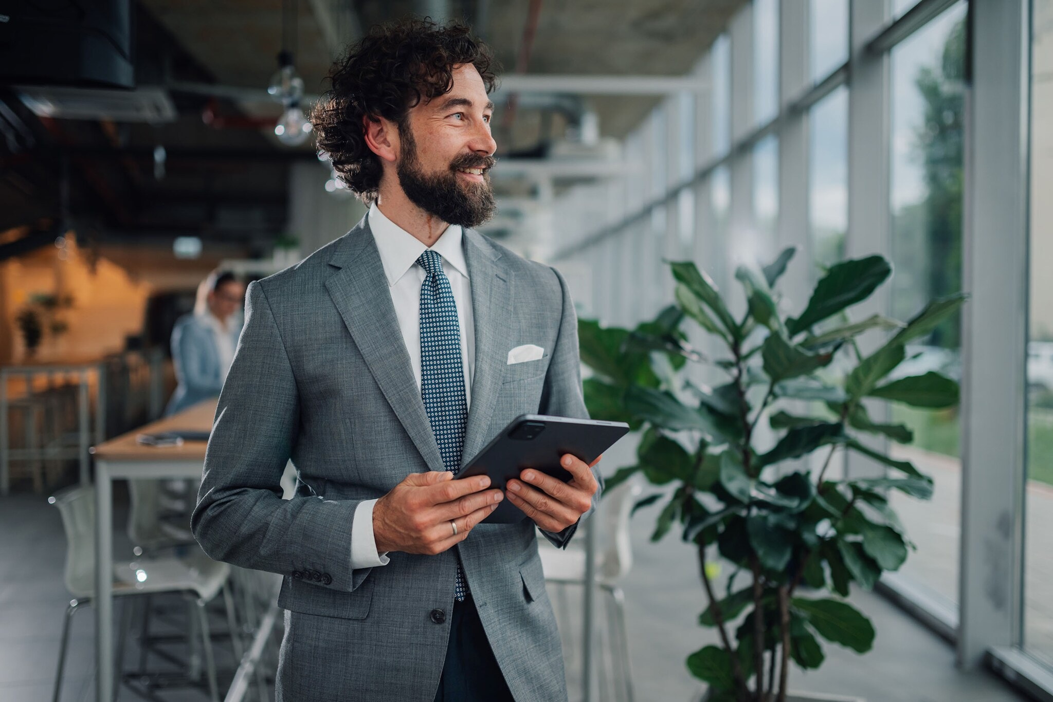Confident businessman holding tablet and smiling in modern office Halbporträt eines lächelnden Mannes im grauen Anzug mit Einstecktuch und Krawatte, der mit einem Tablet in einem Büro steht.