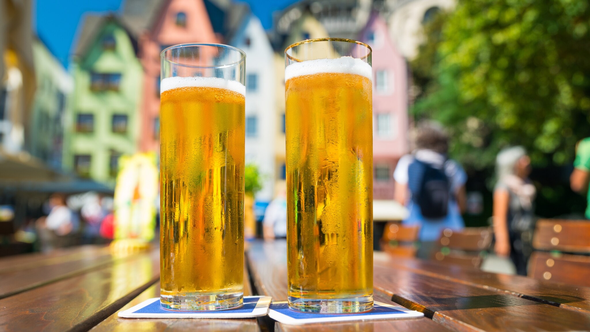 Zwei Gläser Bier auf einem Holztisch, im Hintergrund farbenfrohe Gebäude und Bäume. Die Gläser sind transparent mit goldenem Bier und Schaumkrone. Zwei Gläser Bier auf einem Holztisch, im Hintergrund farbenfrohe Gebäude und Bäume. Die Gläser sind transparent mit goldenem Bier und Schaumkrone.