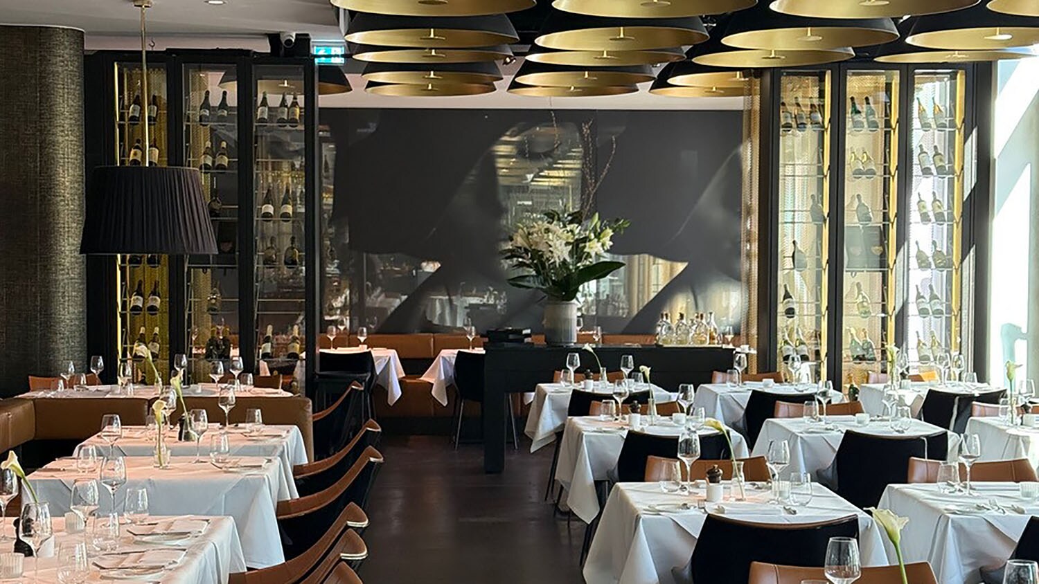 Eleganter Speiseraum mit weiß eingedeckten Tischen und goldfarbenen Deckenlampen vor einem schwarzen Wandbild.