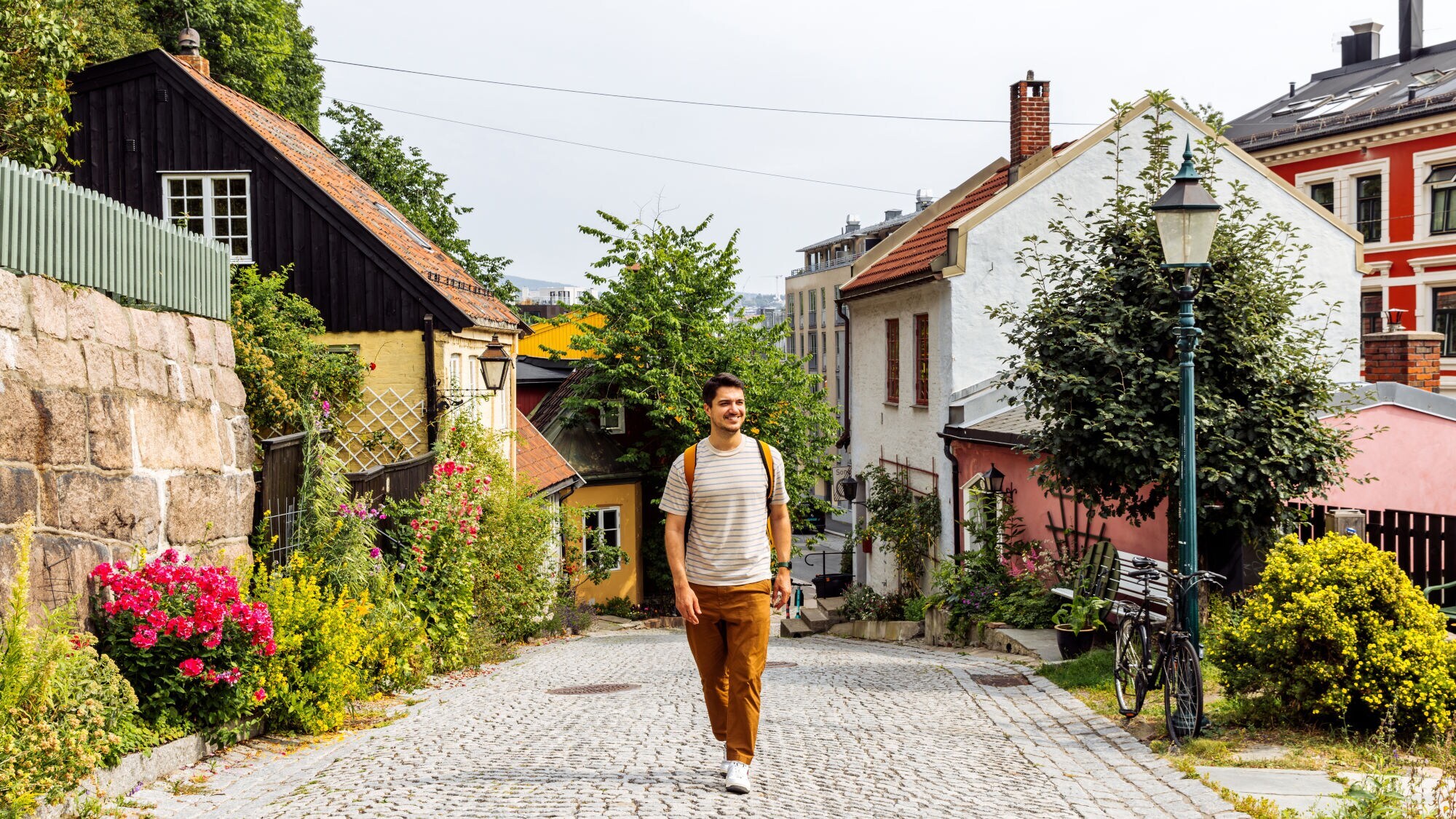 Person mit gestreiftem T-Shirt und brauner Hose geht auf gepflasterter Straße in Oslo, umgeben von bunten Häusern und Blumen.