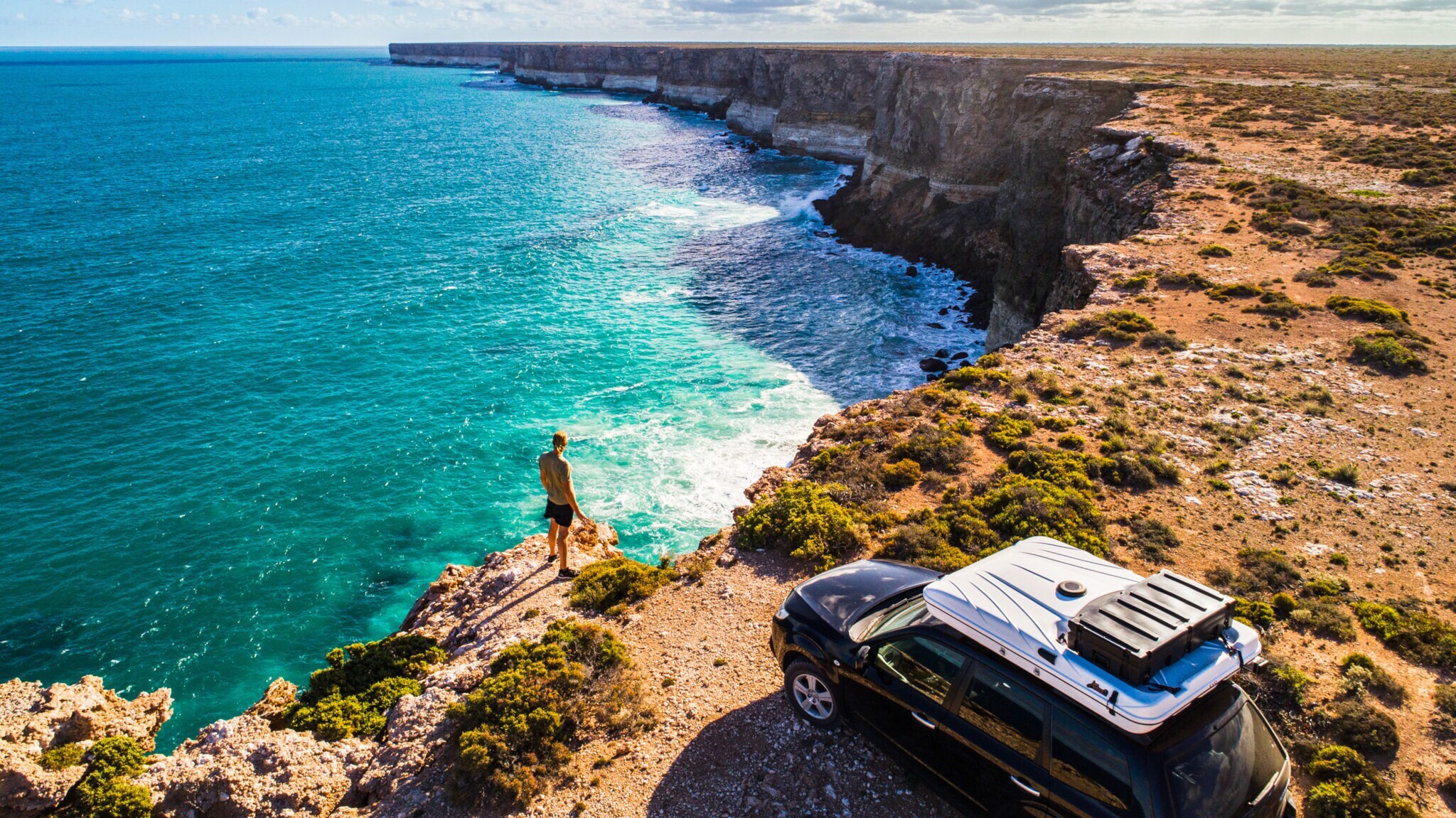 Eine Person steht neben einem Auto mit eingeklappten Dachzelt an einer Felsküste vor türkisblauem Wasser bei Sonnenschein. Eine Person steht neben einem Auto mit eingeklappten Dachzelt an einer Felsküste vor türkisblauem Wasser bei Sonnenschein.