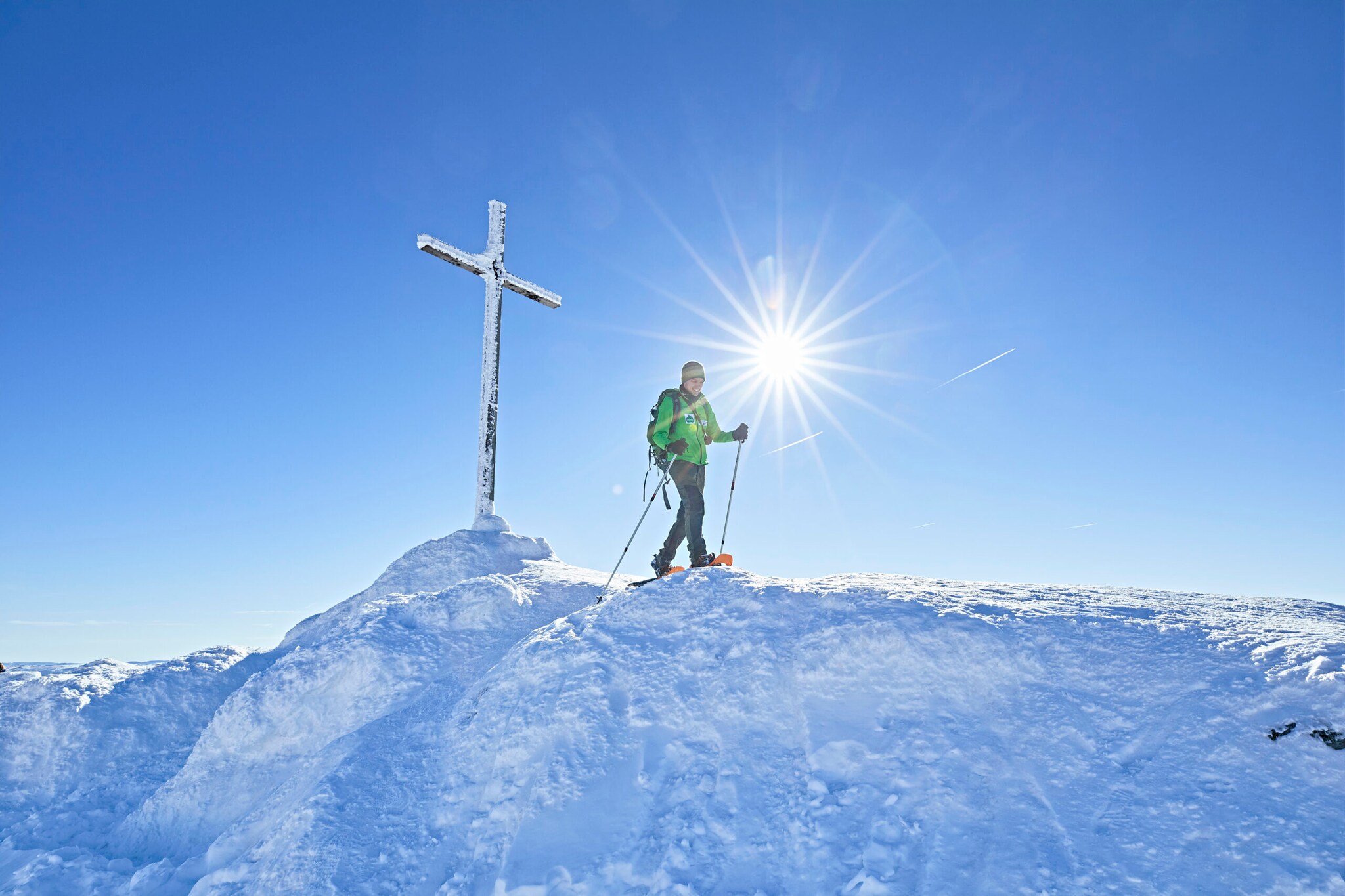 Ein Wanderer mit Schneeschuhen und Stöcken läuft neben einem Gipfelkreuz im Schnee