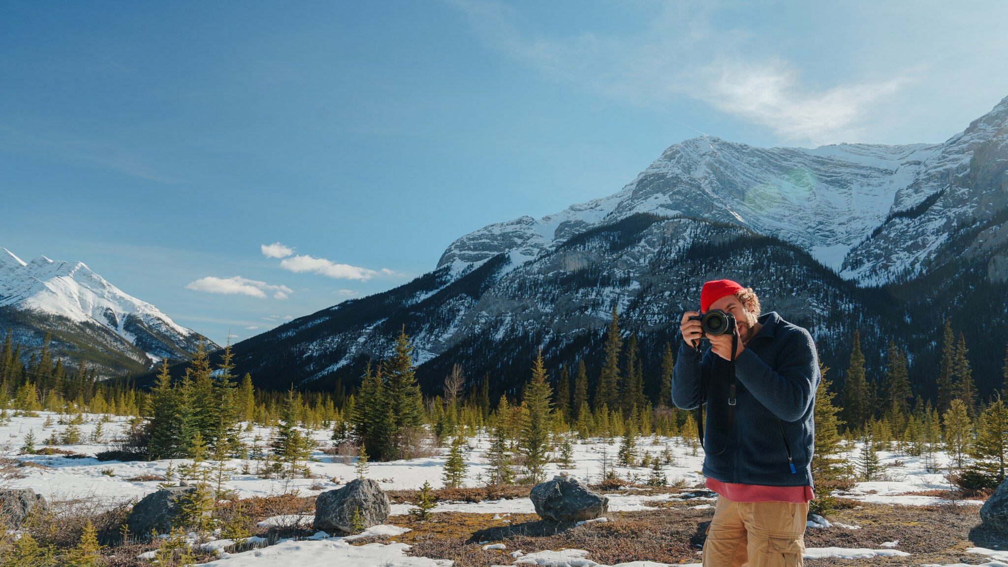 Mann mit roter Mütze und blauer Jacke fotografiert in schneebedeckter Berglandschaft mit Nadelbäumen und schneebedeckten Gipfeln.