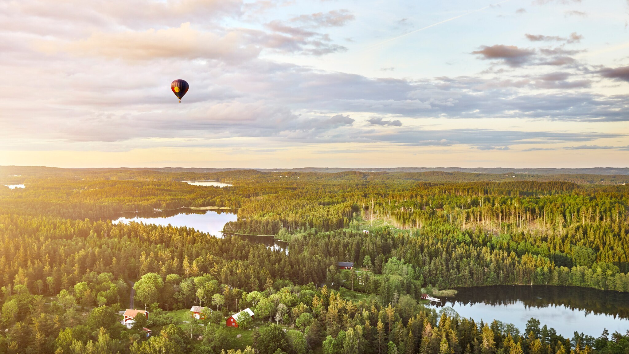 Heißluftballon mit rotem Muster fliegt über bewaldete Landschaft mit mehreren Seen und vereinzelten Häusern bei Sonnenuntergang. Heißluftballon mit rotem Muster fliegt über bewaldete Landschaft mit mehreren Seen und vereinzelten Häusern bei Sonnenuntergang.