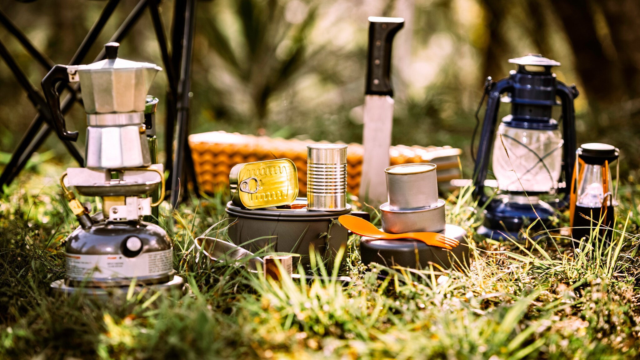 Camping-Kochutensilien auf Gras, darunter Gaskocher mit Espressokocher, Konservendosen, Messer, Pfanne, Laternen und Korb im Hintergrund.