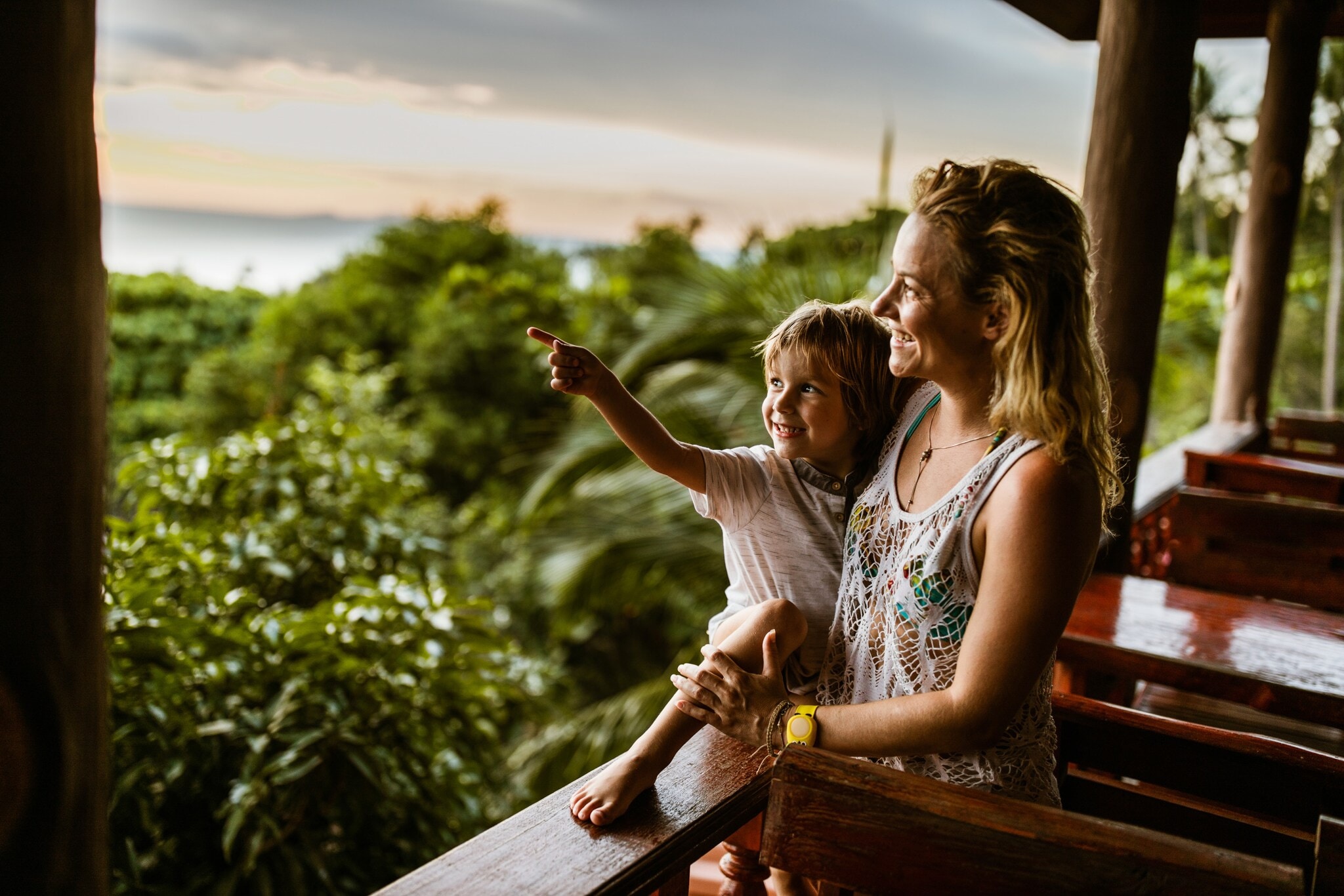 Eine Frau hat einen kleinen Jungen im Arm, beide blicken von einer Holzterrasse in den Dschungel von Thailand, hinter ihnen das Meer. Eine Frau hat einen kleinen Jungen im Arm, beide blicken von einer Holzterrasse in den Dschungel von Thailand, hinter ihnen das Meer.