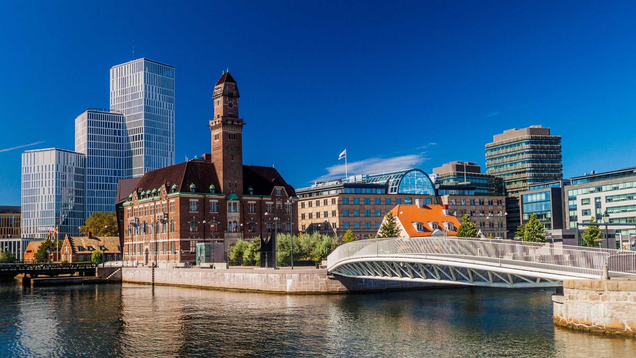 Blick auf einen Fluss mit einer modernen Fußgängerbrücke und historischen sowie modernen Gebäuden im Hintergrund unter blauem Himmel. Blick auf einen Fluss mit einer modernen Fußgängerbrücke und historischen sowie modernen Gebäuden im Hintergrund unter blauem Himmel.
