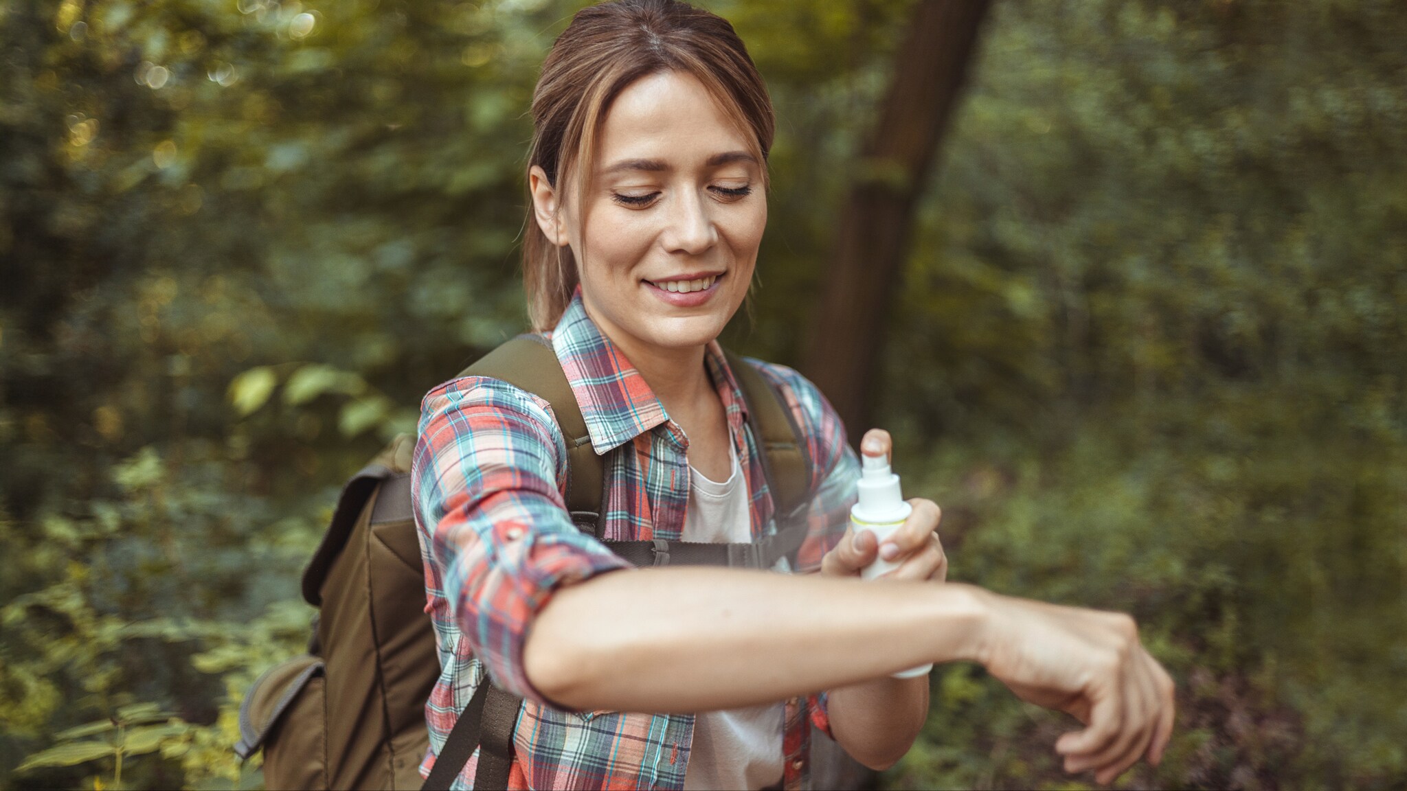 Person mit kariertem Hemd und Rucksack trägt im Wald Insektenschutzspray auf den Arm auf.