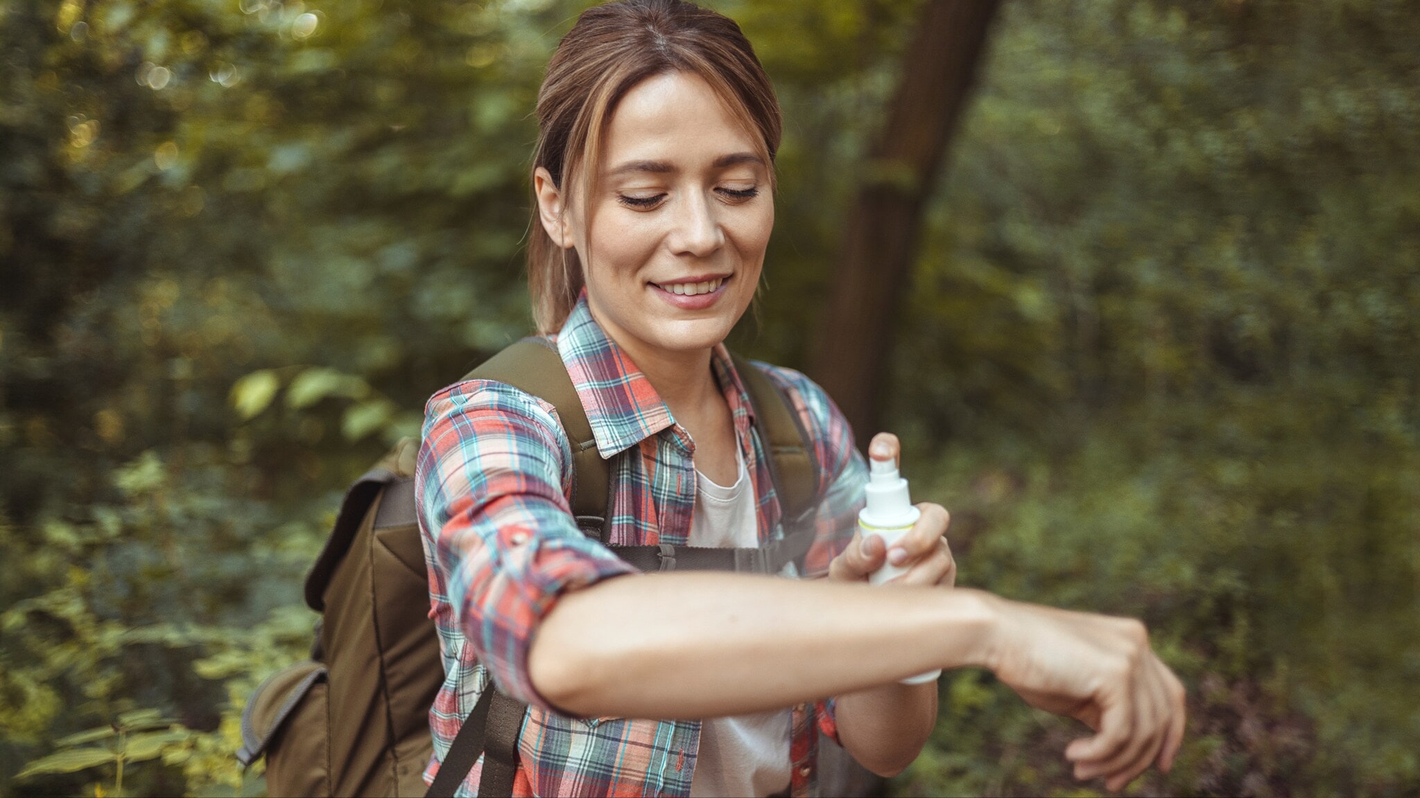 Person mit kariertem Hemd und Rucksack trägt im Wald Insektenschutzspray auf den Arm auf. Person mit kariertem Hemd und Rucksack trägt im Wald Insektenschutzspray auf den Arm auf.