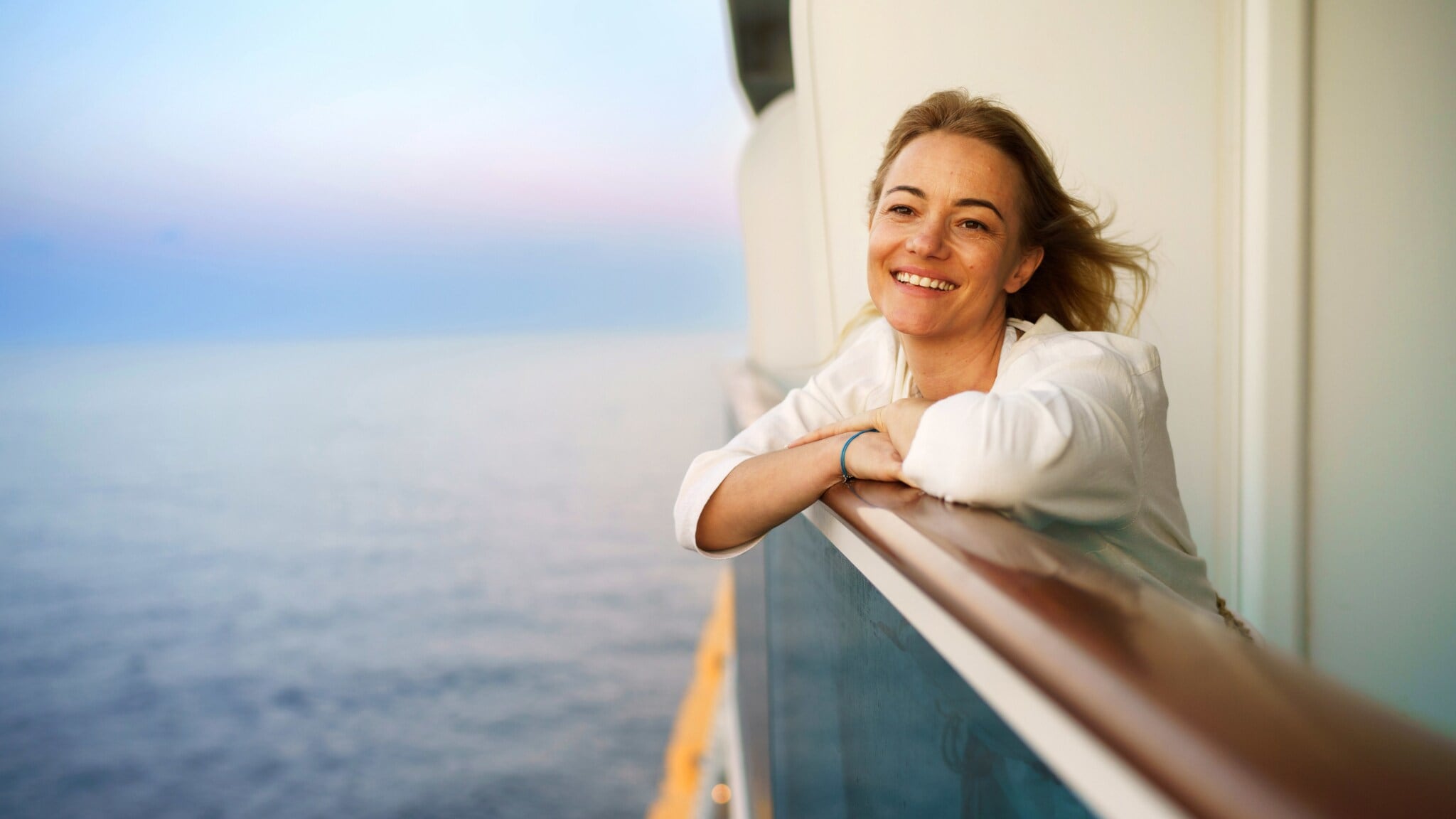 Eine lächelnde Frau lehnt an der Reling einer Balkonkabine auf einem Kreuzfahrtschiff auf See bei Abenddämmerung. Eine lächelnde Frau lehnt an der Reling einer Balkonkabine auf einem Kreuzfahrtschiff auf See bei Abenddämmerung.