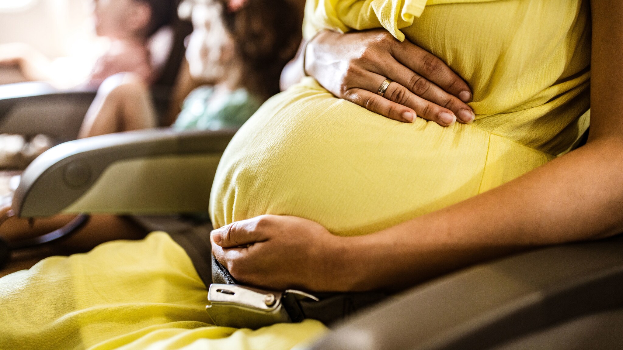Nahaufnahme eines Schwangerschaftsbauch, umfasst von zwei Händen einer Frau im gelben Kleid auf einem Flugzeugsitz neben zwei Kindern.