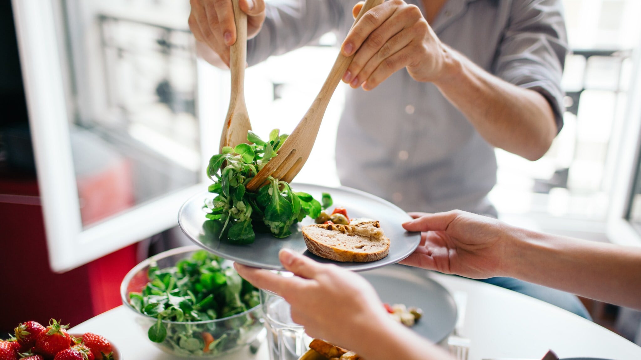 Personen servieren frischen grünen Salat mit Holzbesteck auf Teller mit Brot und Gemüse, auf dem Tisch Schüssel mit Salat und Schale mit Erdbeeren. Personen servieren frischen grünen Salat mit Holzbesteck auf Teller mit Brot und Gemüse, auf dem Tisch Schüssel mit Salat und Schale mit Erdbeeren.