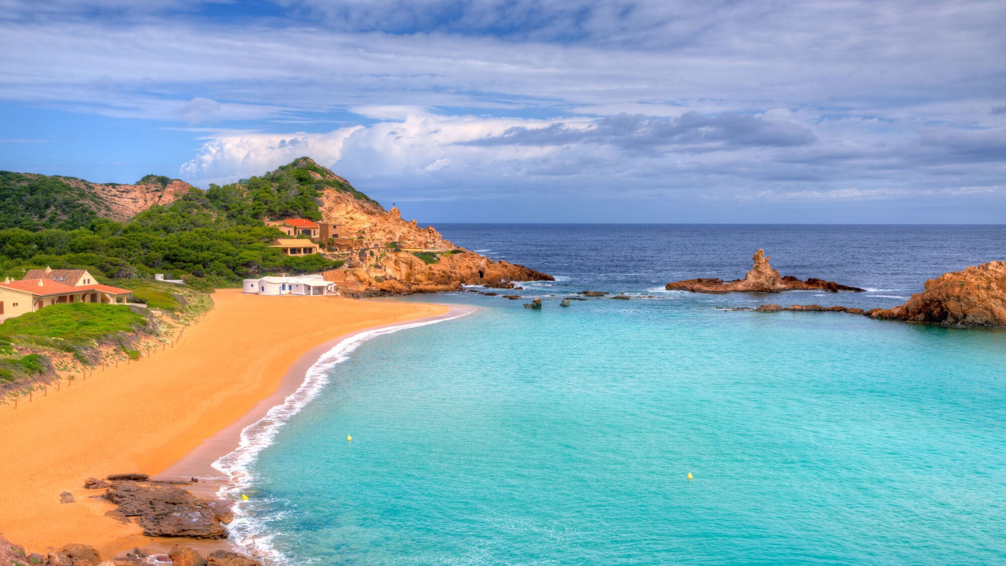 Strand mit feinem Sand und klarem Wasser, umgeben von grünen Hügeln und Felsen. Im Hintergrund sind einige Häuser sichtbar.
