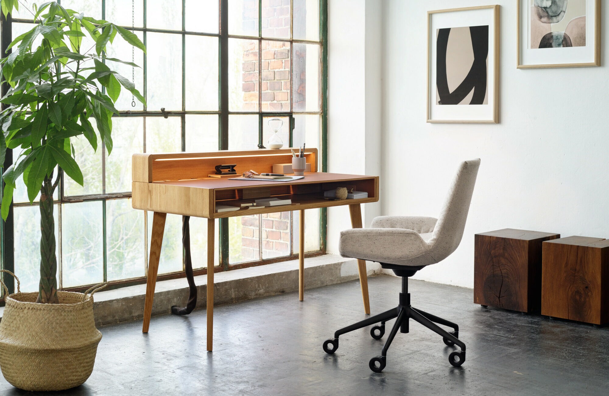 Ein Sekretär aus Holz und ein gepolsterter Schreibtischstuhl auf Rollen stehen in einem modern eingerichteten, hellen Raum mit bodentiefen Fenstern mit Loft-Charakter