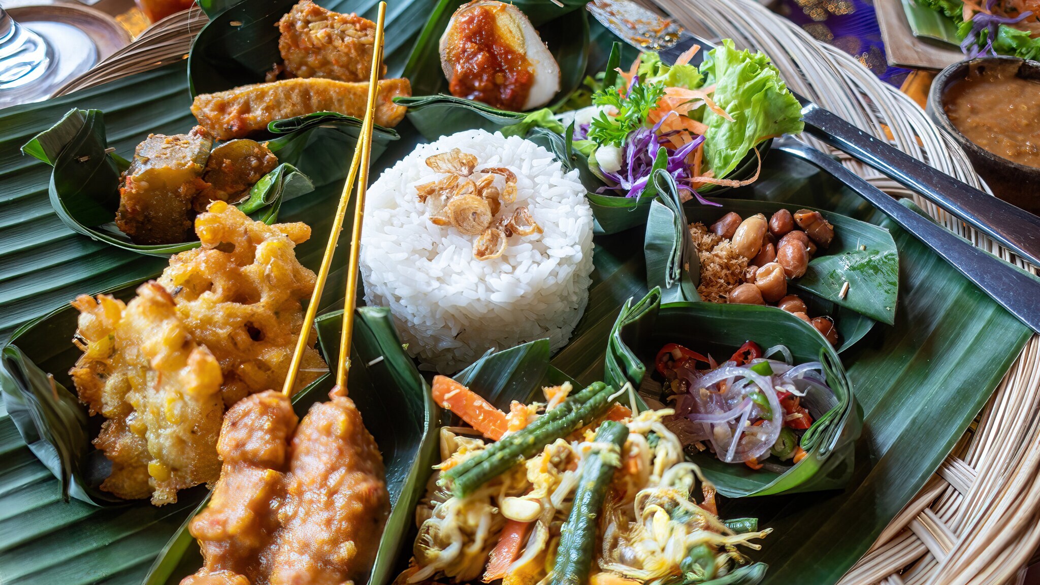in runder Korb mit verschiedenen indonesischen Gerichten, darunter Reis, Gemüse, frittiertes Essen und Erdnüsse, serviert auf Bananenblättern.