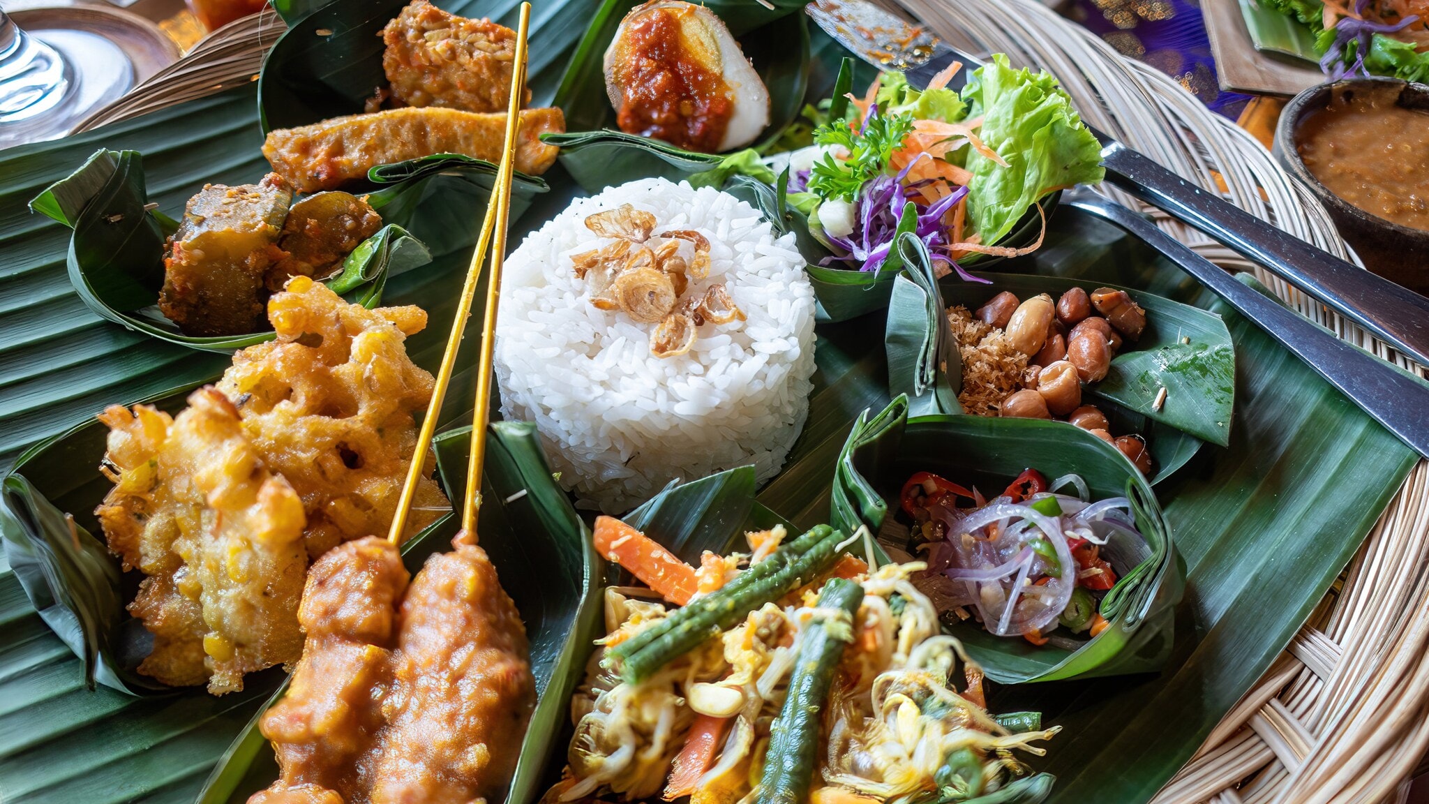 in runder Korb mit verschiedenen indonesischen Gerichten, darunter Reis, Gemüse, frittiertes Essen und Erdnüsse, serviert auf Bananenblättern. in runder Korb mit verschiedenen indonesischen Gerichten, darunter Reis, Gemüse, frittiertes Essen und Erdnüsse, serviert auf Bananenblättern.