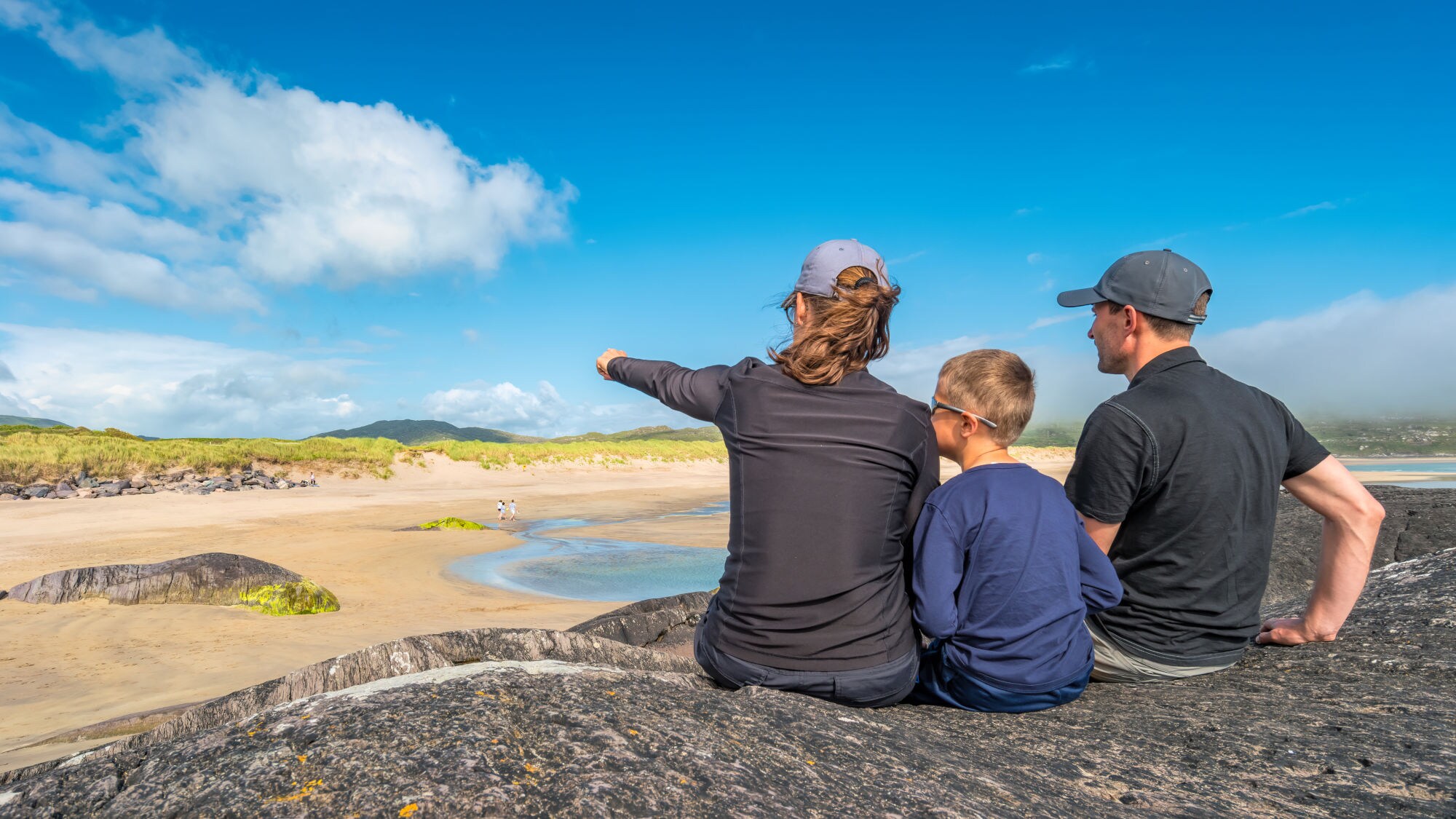 Drei Personen sitzen auf einem Felsen und blicken auf einen Sandstrand mit Dünen und blauem Himmel in Irland.