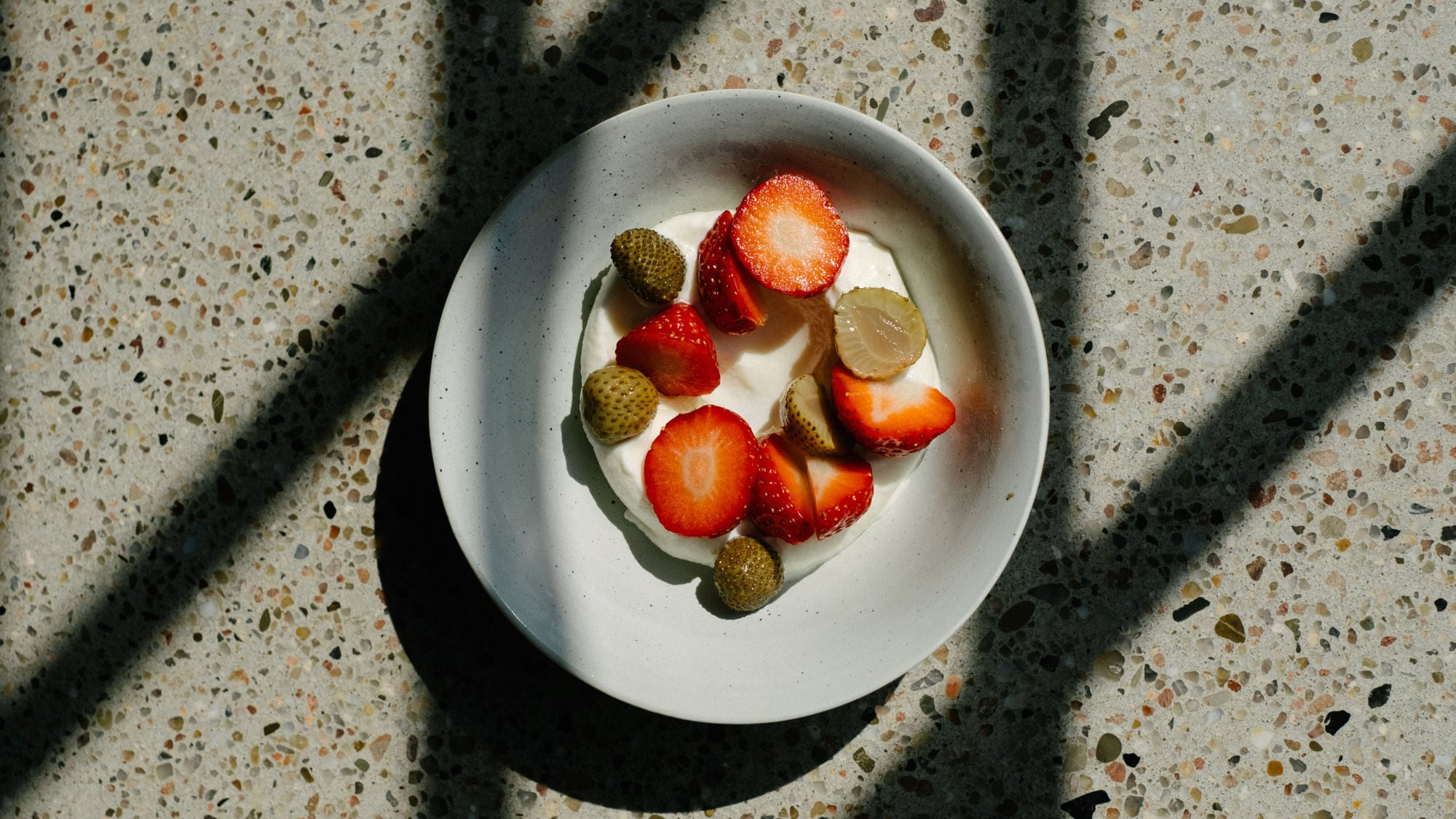 Schale mit Joghurt, belegt mit geschnittenen Erdbeeren und kleinen grünen Früchten, auf marmoriertem Untergrund mit Schatten. Schale mit Joghurt, belegt mit geschnittenen Erdbeeren und kleinen grünen Früchten, auf marmoriertem Untergrund mit Schatten.