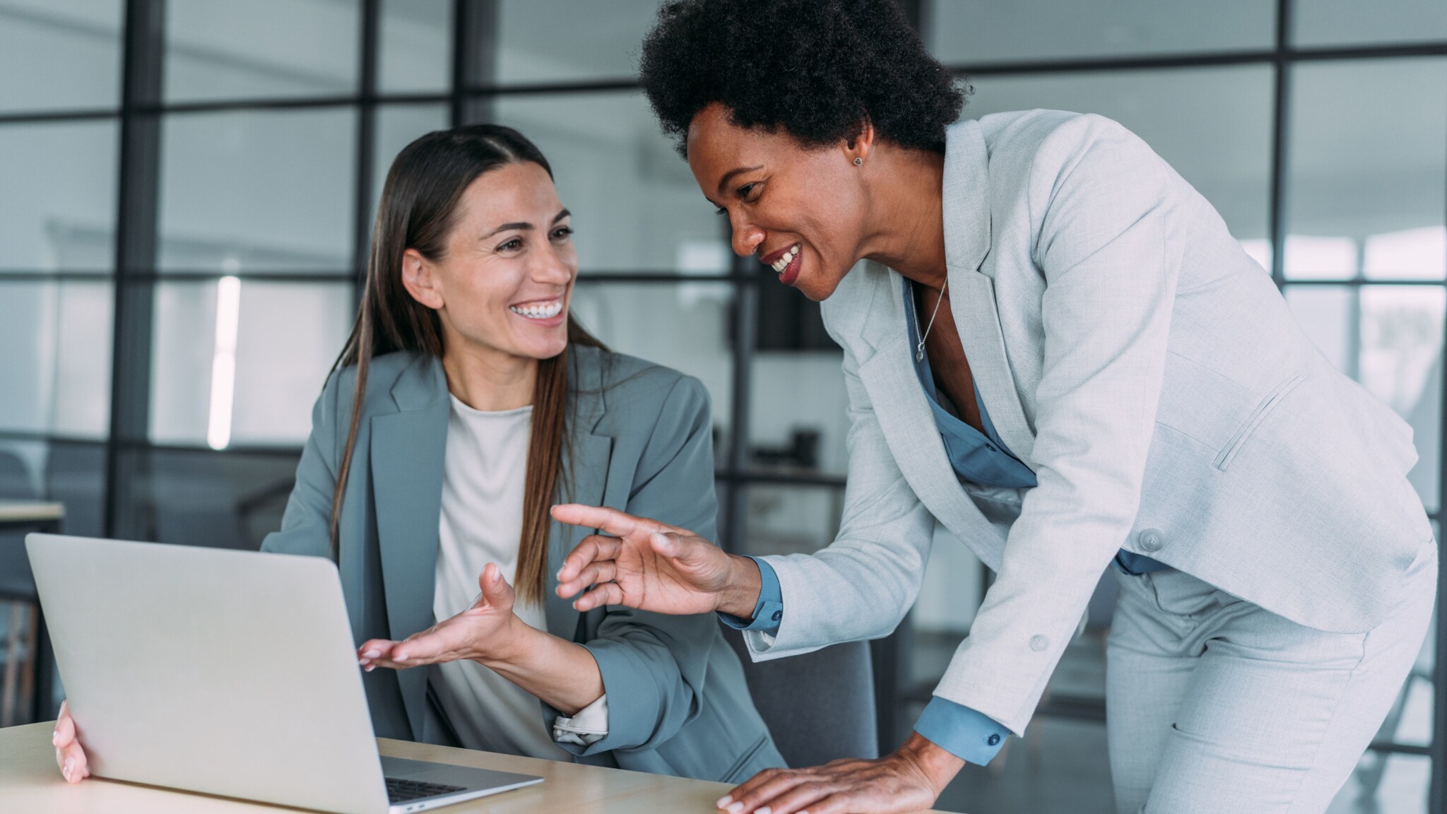 Zwei Frauen in grauen Blazern kommunizieren lächelnd an einem Arbeitsplatz mit Laptop.