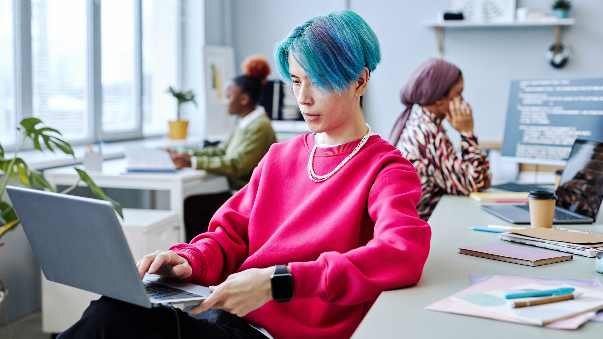 Ein junger Mann mit blauen Haaren im pinkfarbenen Pullover sitzt mit Laptop auf dem Schoß in einem Büro, im Hintergrund zwei Frauen an weiteren Arbeitsplätzen.