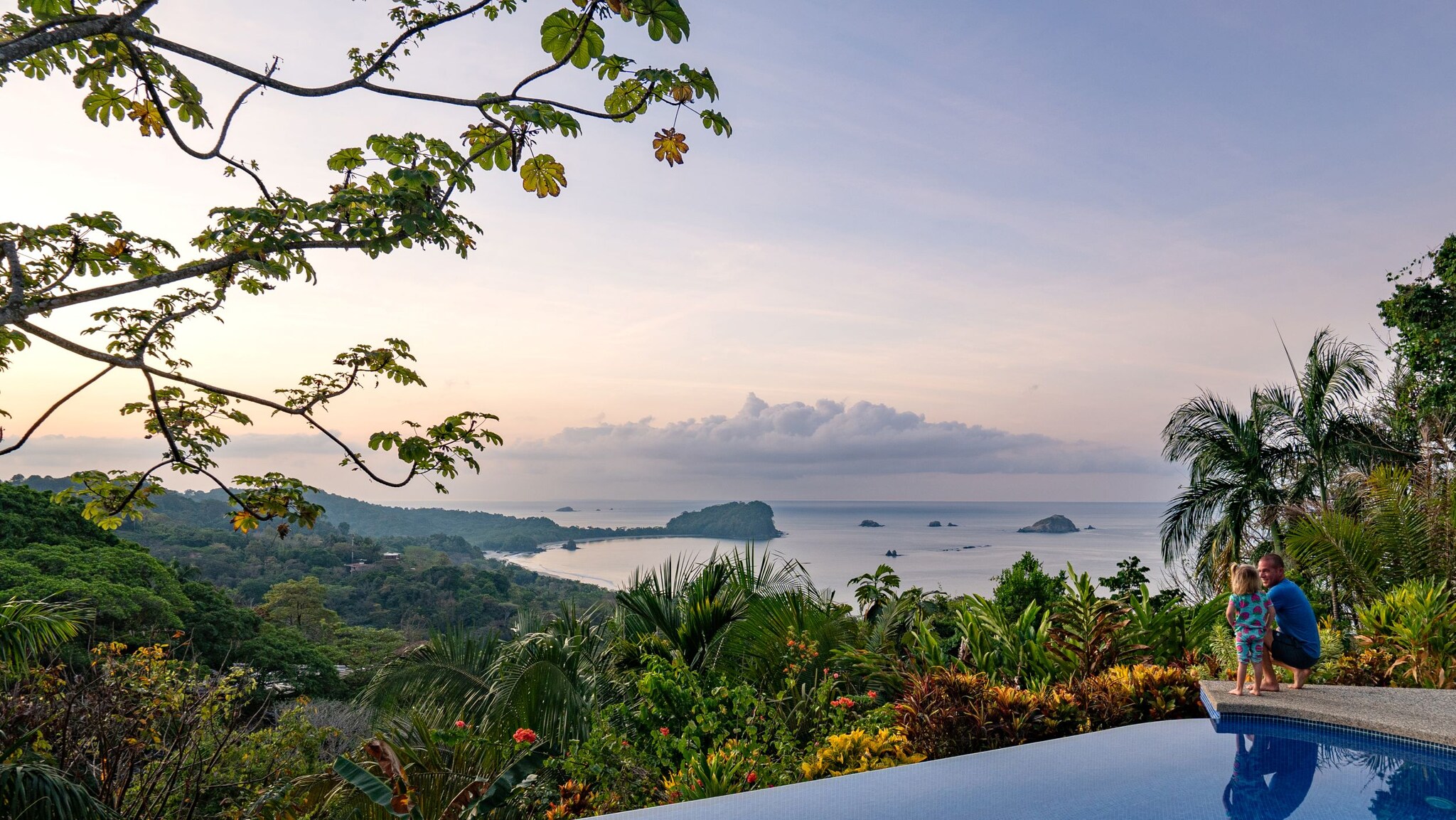 Blick auf tropische Küstenlandschaft mit Inseln im Meer, Pflanzen und Infinity-Pool im Vordergrund, zwei Personen am Poolrand. Blick auf tropische Küstenlandschaft mit Inseln im Meer, Pflanzen und Infinity-Pool im Vordergrund, zwei Personen am Poolrand.
